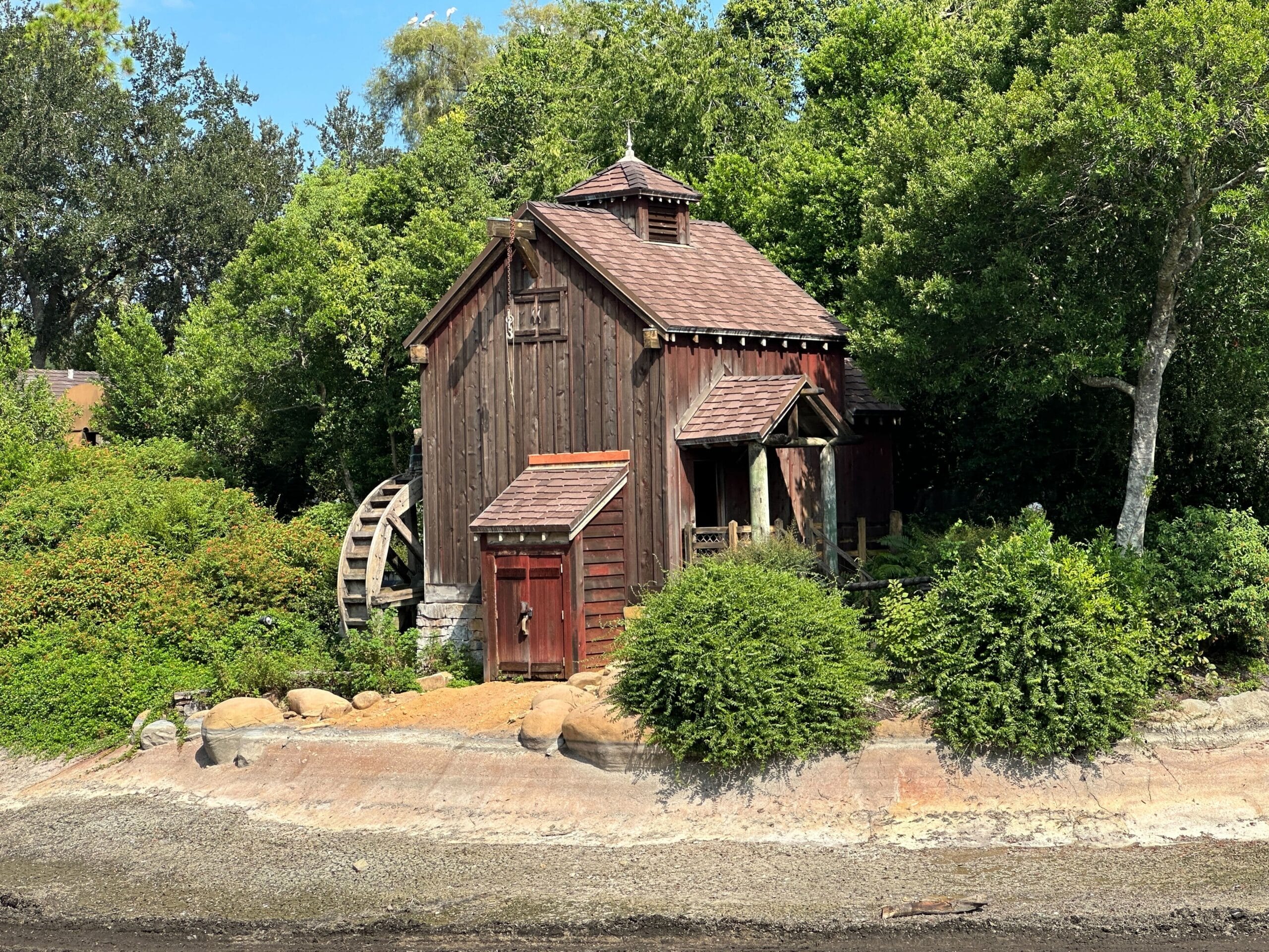 A rustic wooden watermill stands by a dry, rocky riverbank near the Rivers of America, its old track removed.