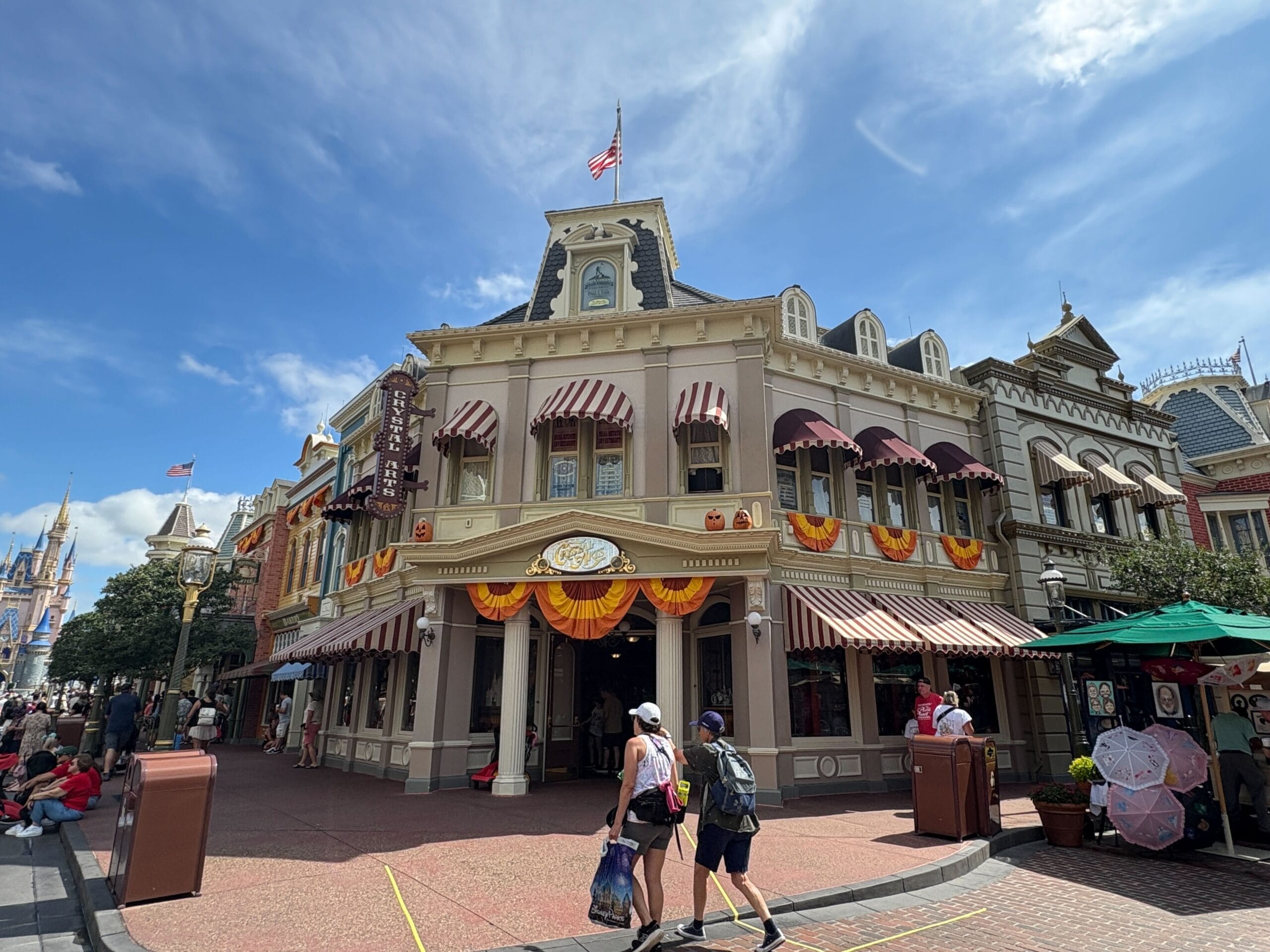 People walk past a bakery with Halloween decor on a sunny day at Magic Kingdom, with the castle in the background.