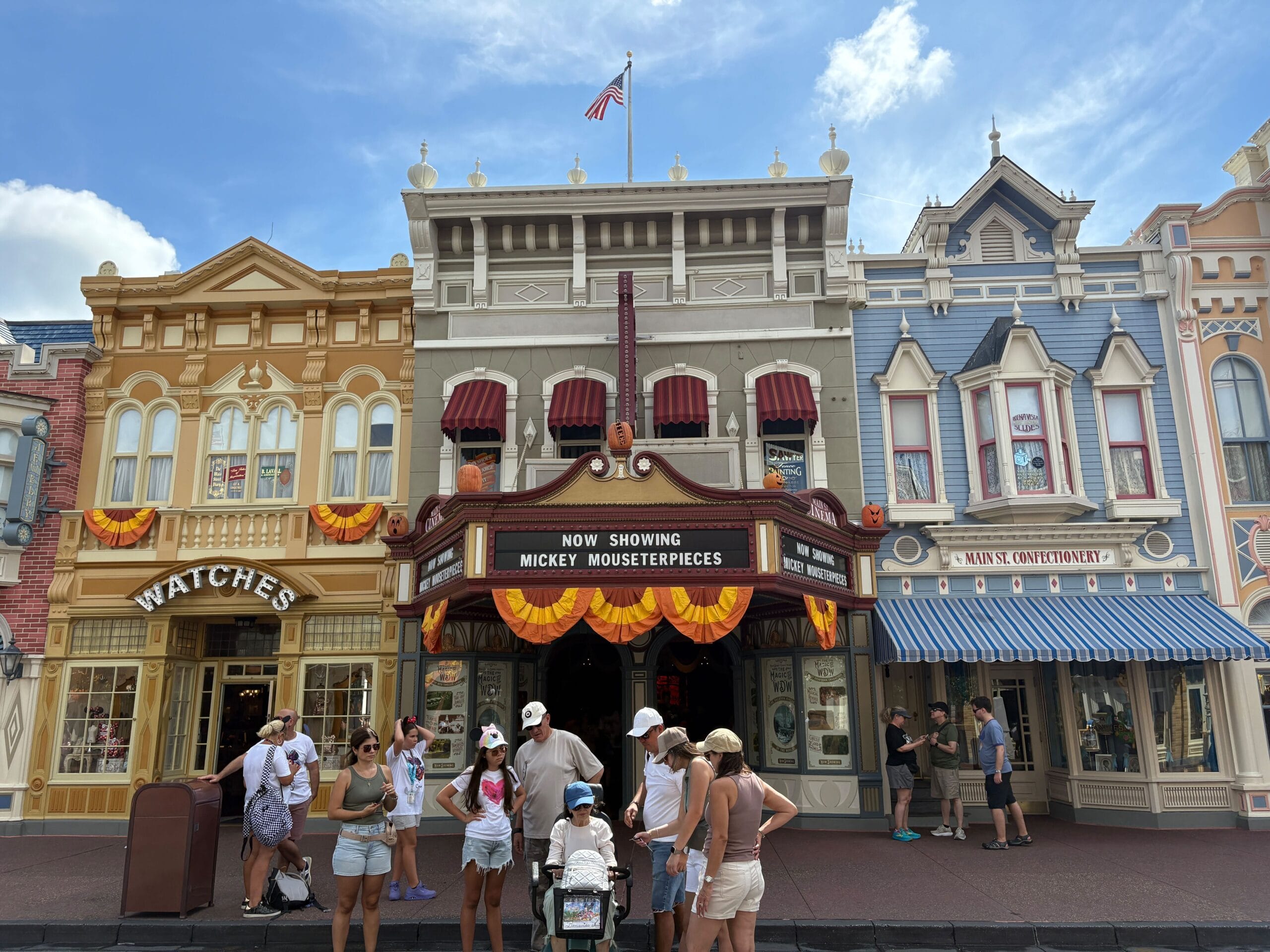 People stand before colorful Magic Kingdom buildings adorned with Halloween decor, near a Mickey Mouse-themed theater sign.