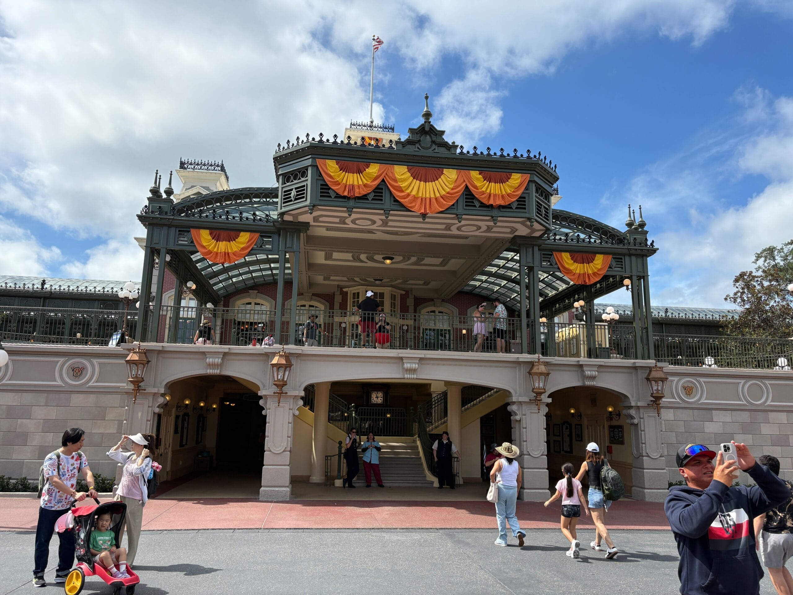 Main entrance to Magic Kingdom decked with Halloween Decor; people stroll beneath orange banners, snapping festive photos.