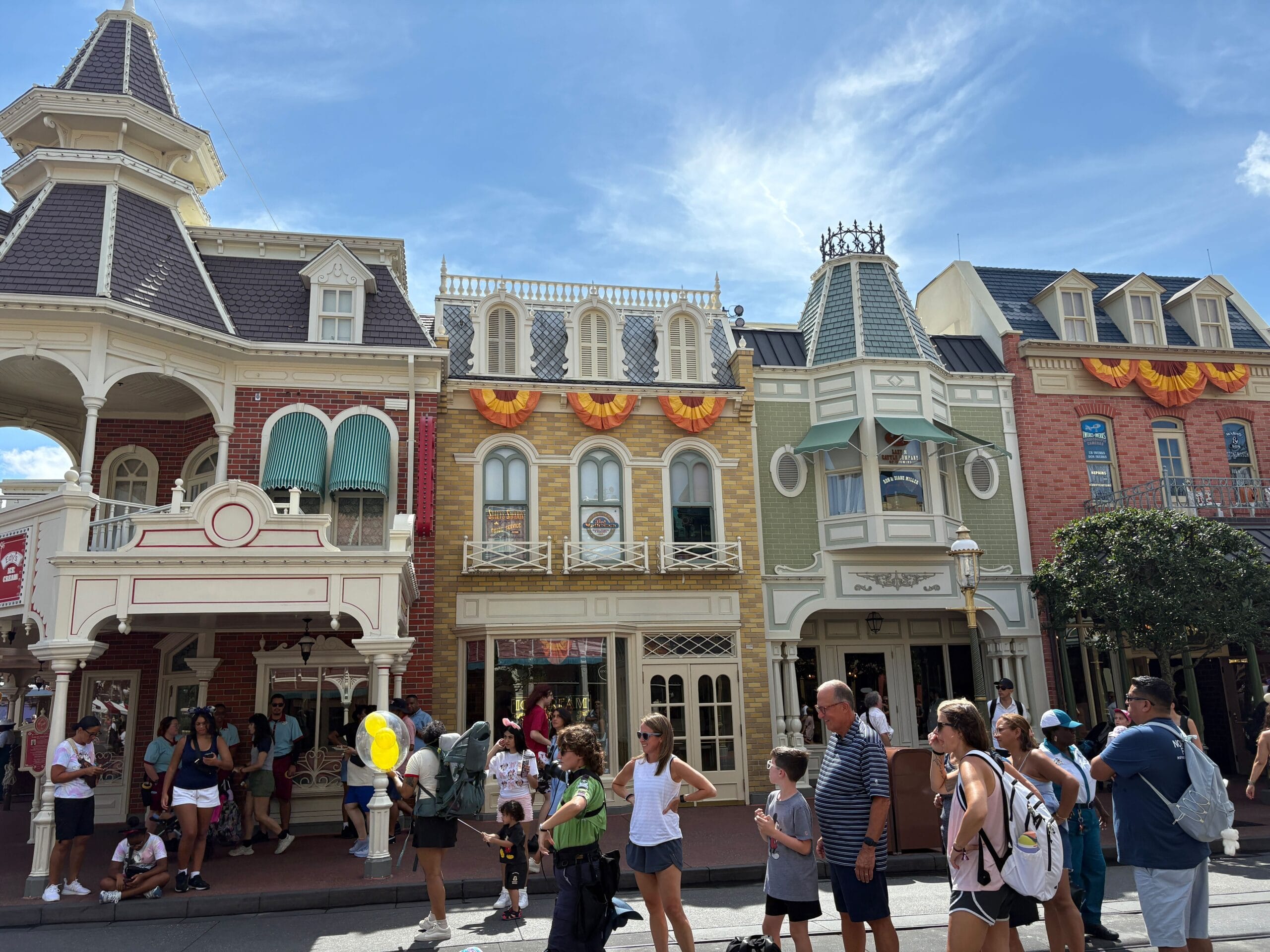 People walk past colorful Main Street buildings adorned with Halloween Decor at Magic Kingdom under a sunny blue sky.