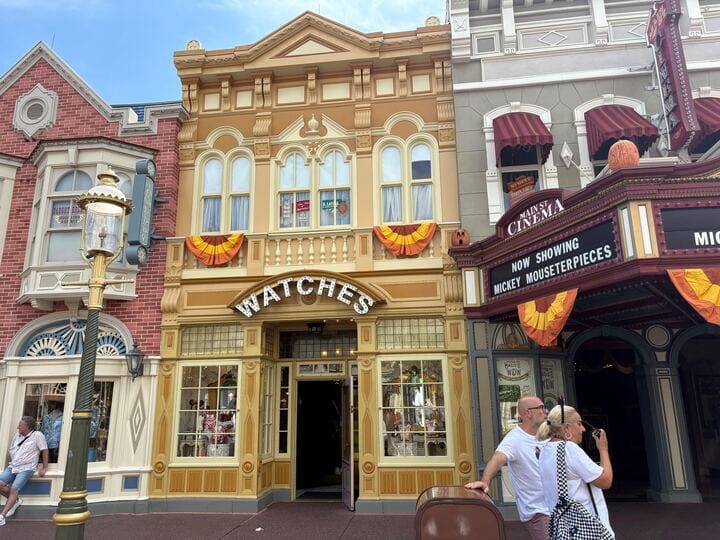Storefront with "Watches" sign, Halloween decor, and two people outside on a sunny day in Magic Kingdom.
