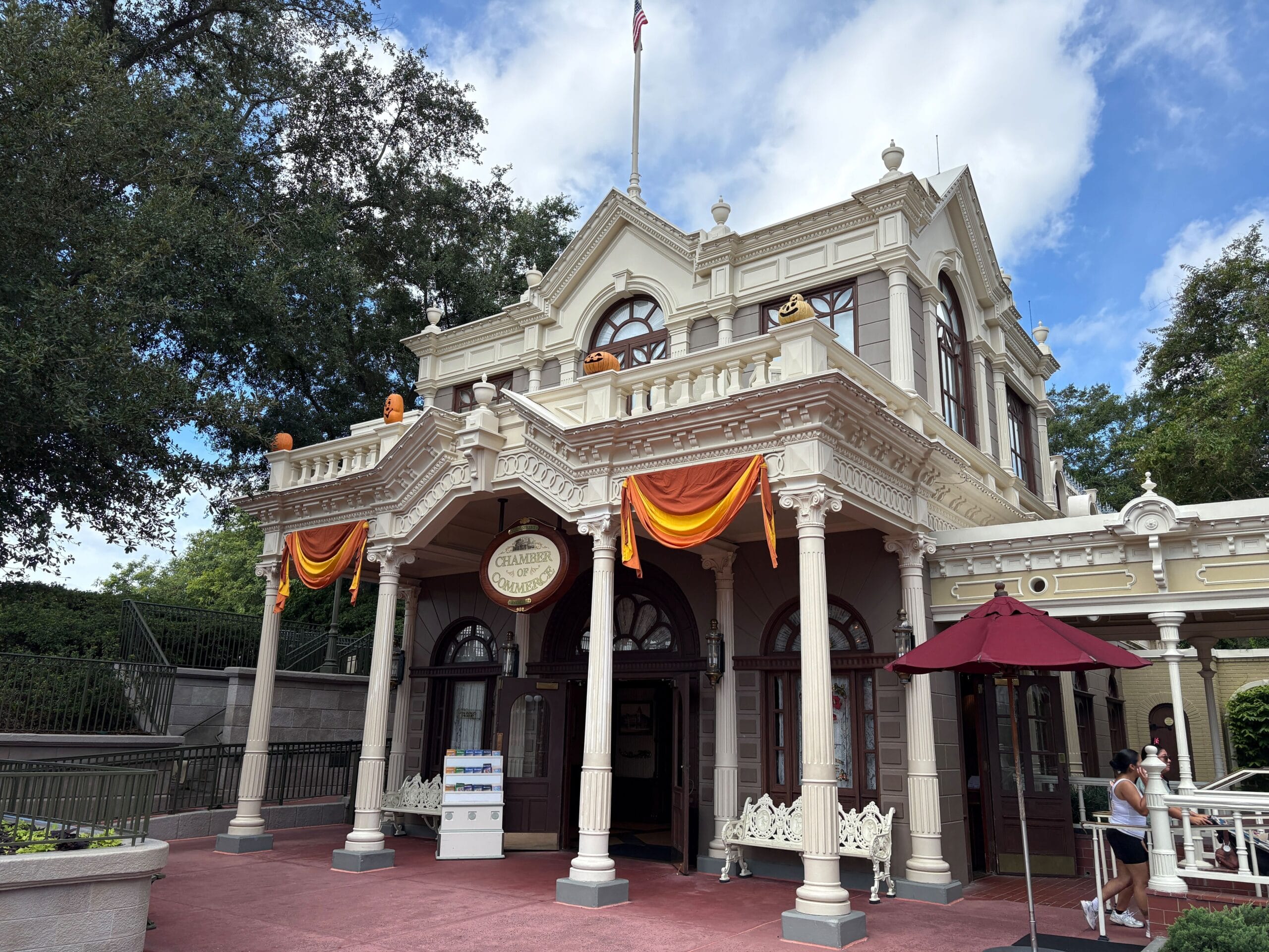 Victorian-style building with Halloween decor at Magic Kingdom, orange banners, pumpkins, and a cloudy blue sky above.