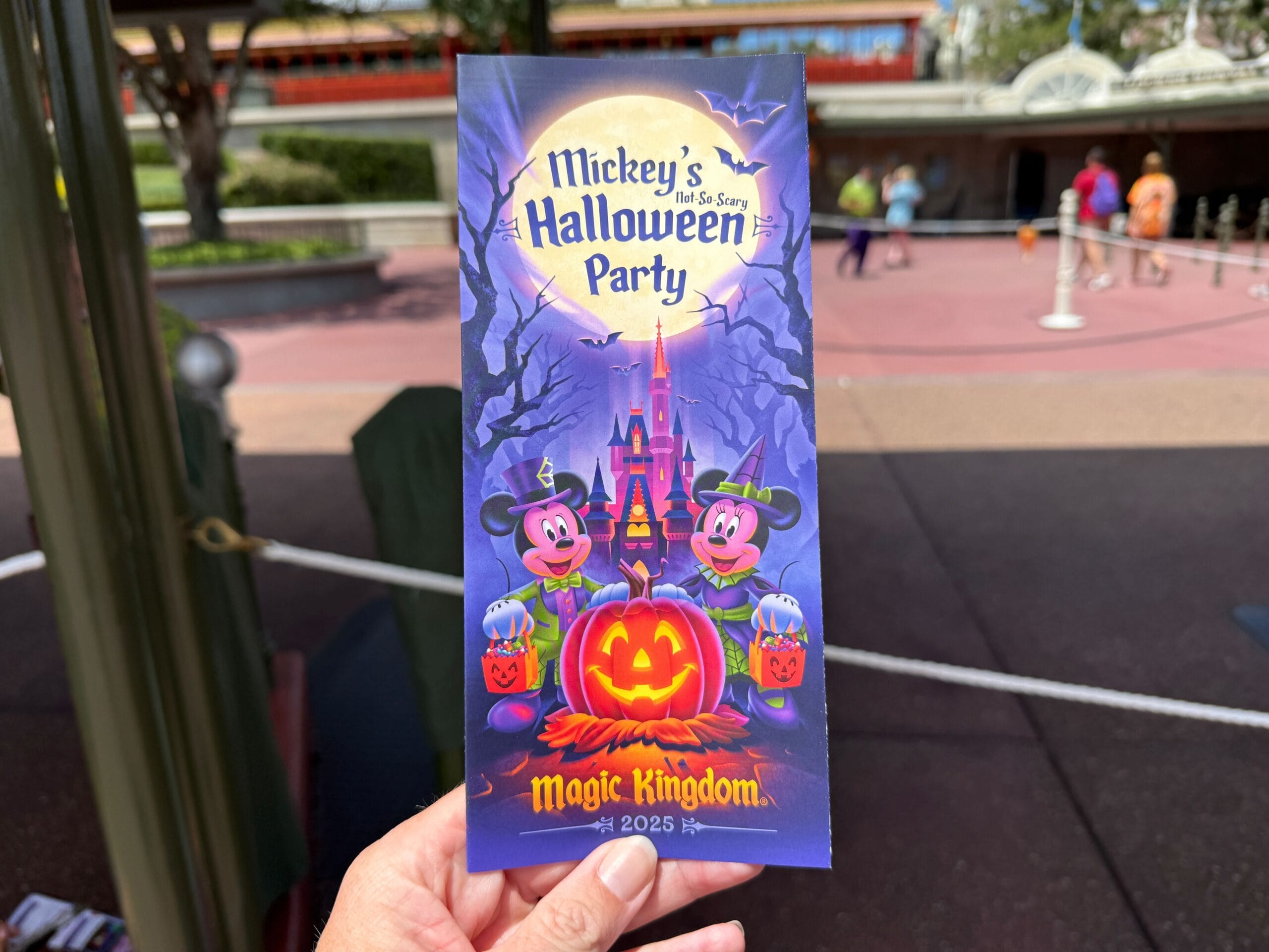 A hand holds a 2025 Mickey's Not So Scary Halloween Party map with Mickey and Minnie in costumes at Magic Kingdom.