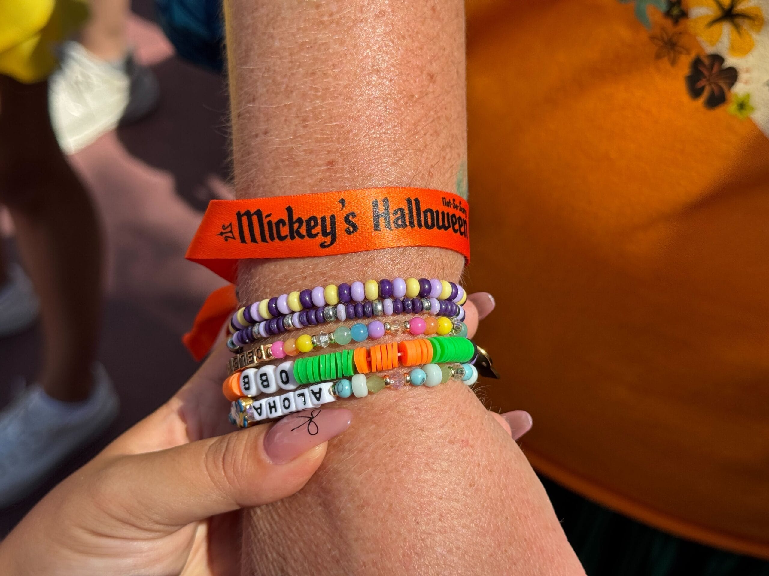 A wrist with colorful beaded bracelets and a 2025 "Mickey's Not So Scary Halloween Party" treat bag, held by another hand.