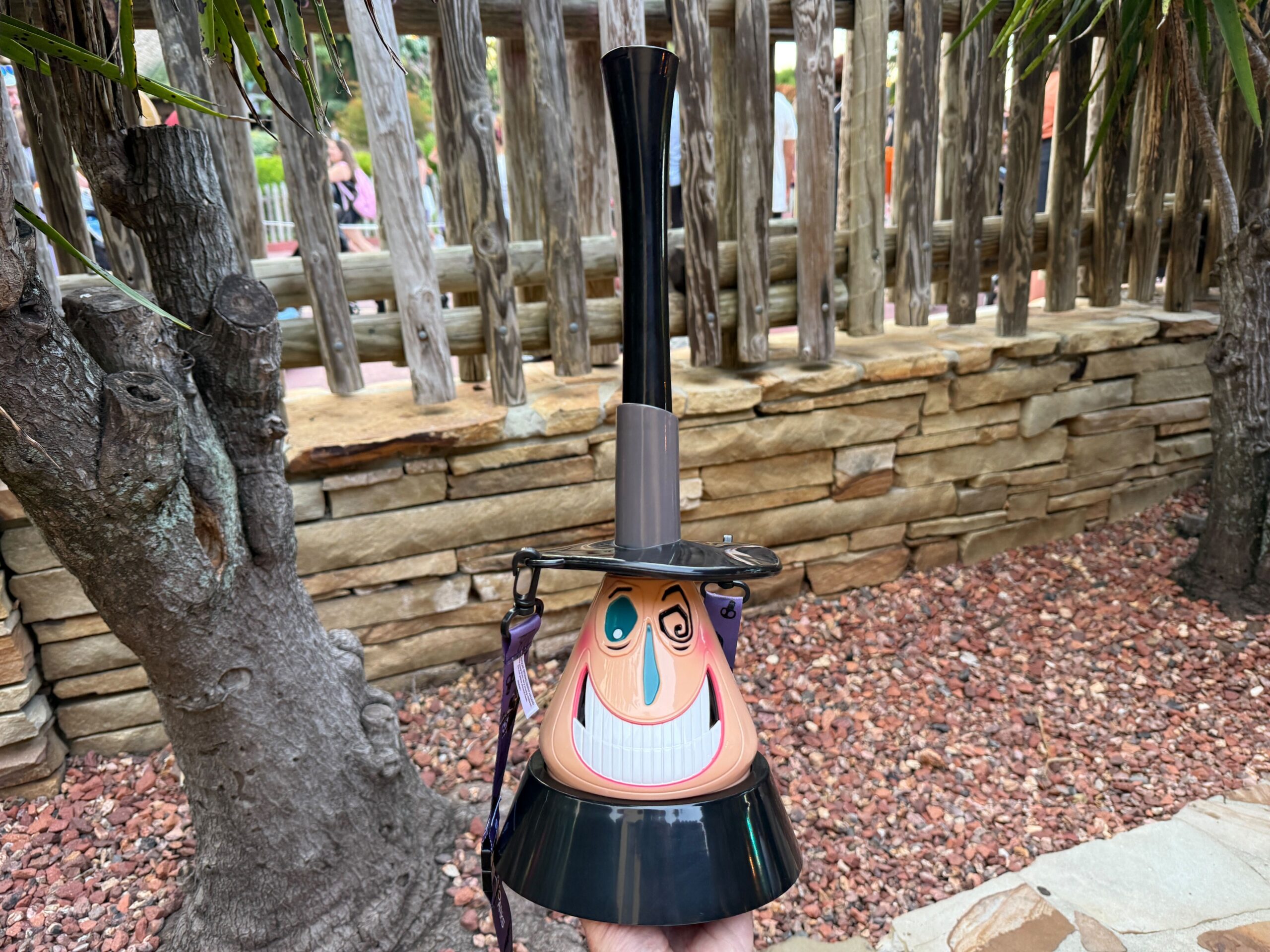 A hand holds a tall Mayor Sipper cup shaped like a smiling cartoon witch, with a stone wall and trees in the background.