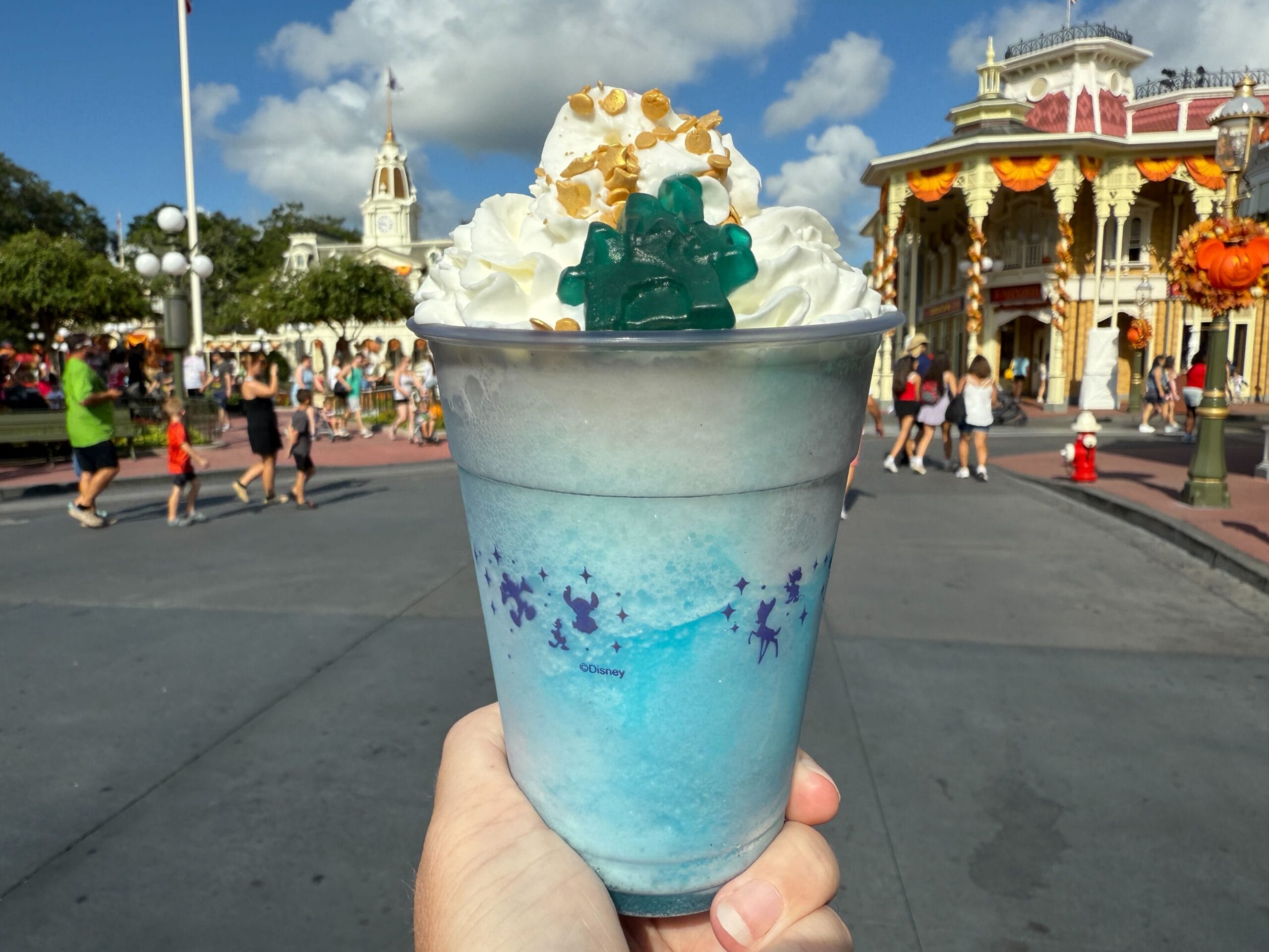 A hand holds a blue frozen drink with whipped cream, sprinkles, and a green gummy, in a theme park decorated for fall.