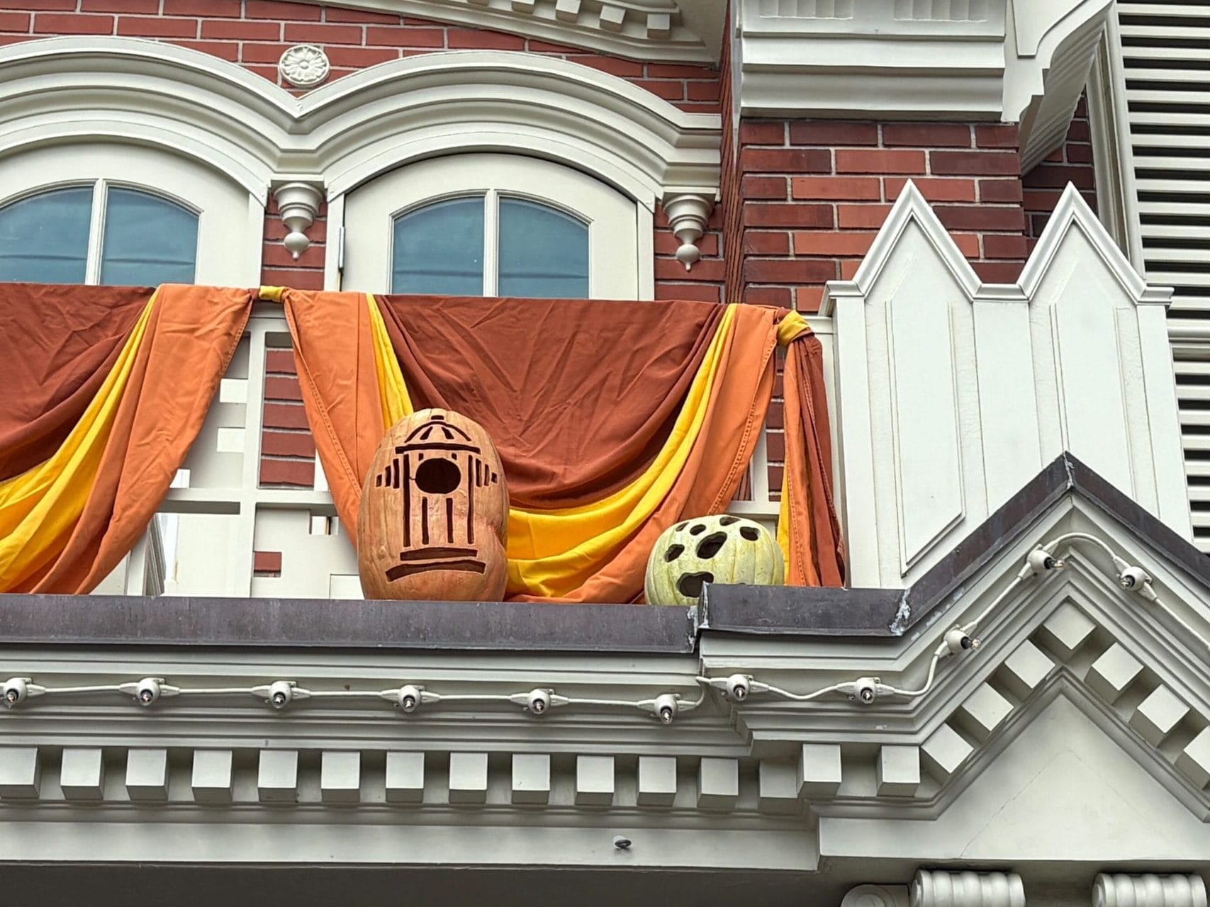 Orange and yellow fabric drapes with Halloween decor and two carved pumpkins on a white balcony of a brick building.
