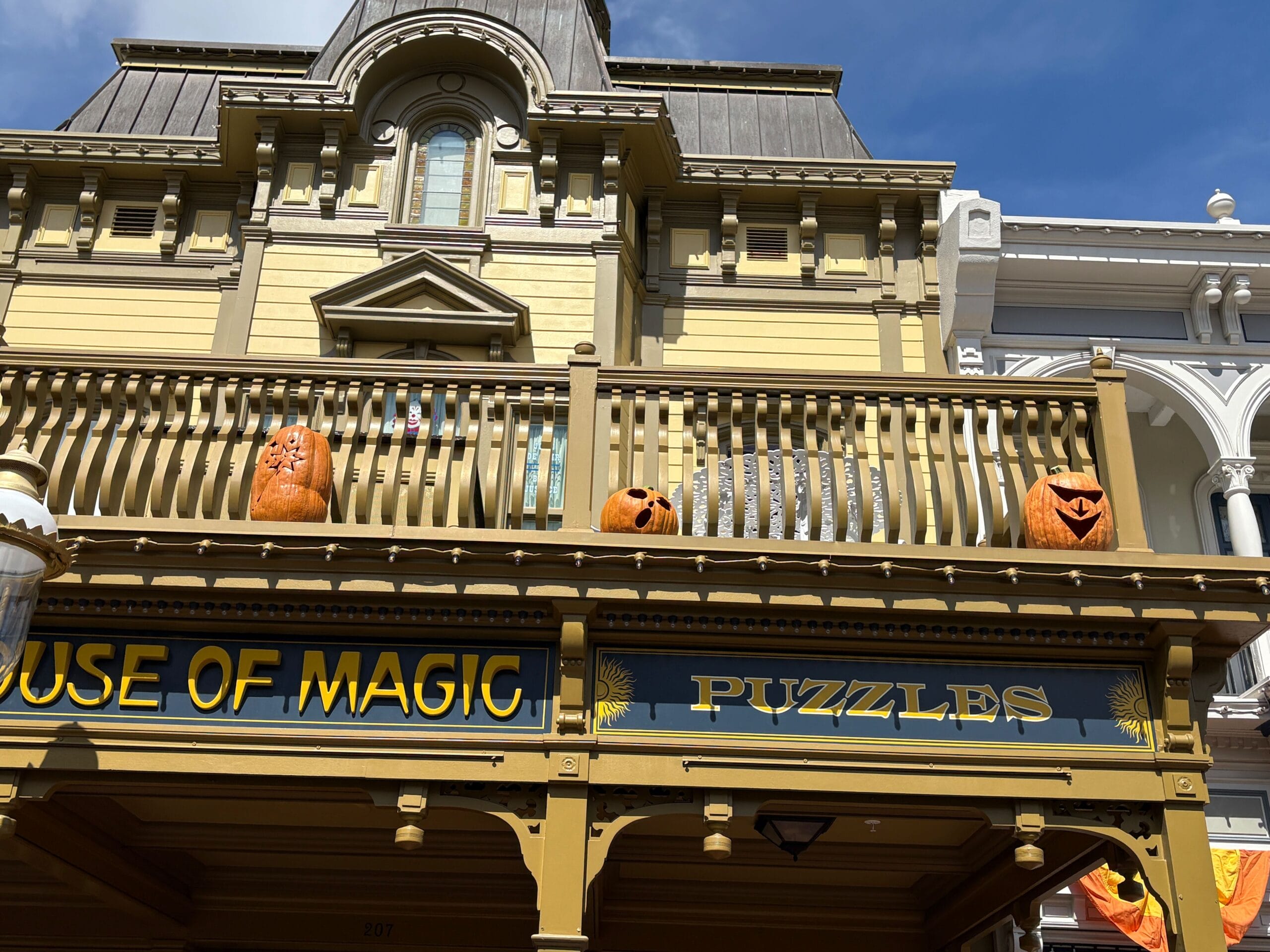 Three carved pumpkins with faces sit as Halloween Decor above a "House of Magic Puzzles" sign on a sunny day.