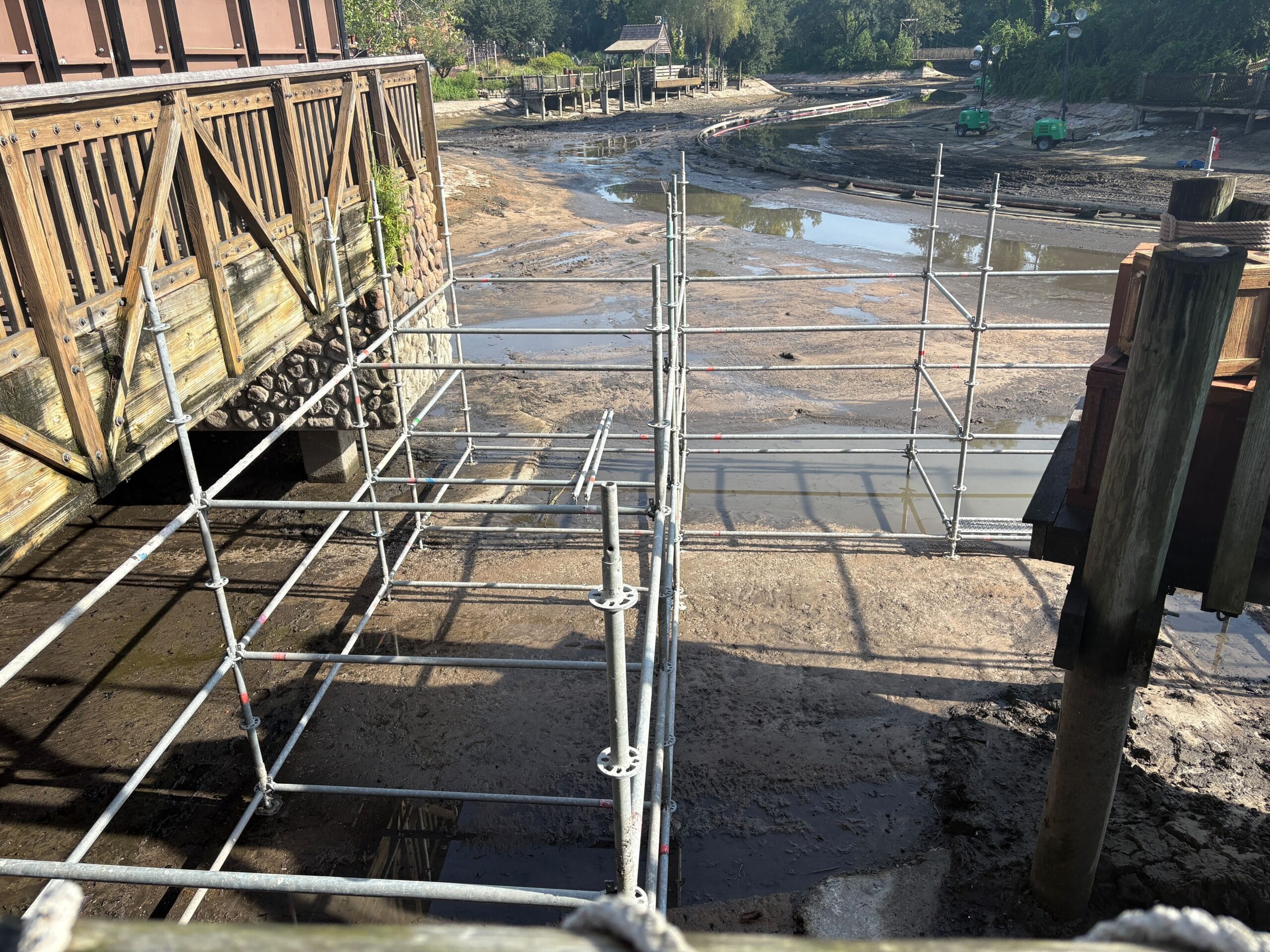 Scaffolding set up over a muddy, drained waterway near a wooden deck and surrounding construction area.