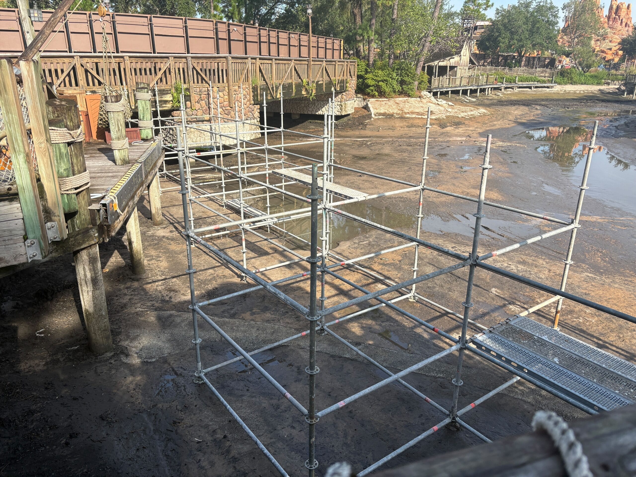 Metal scaffolding stands over a muddy, drained area near a wooden bridge and fence in a park setting.