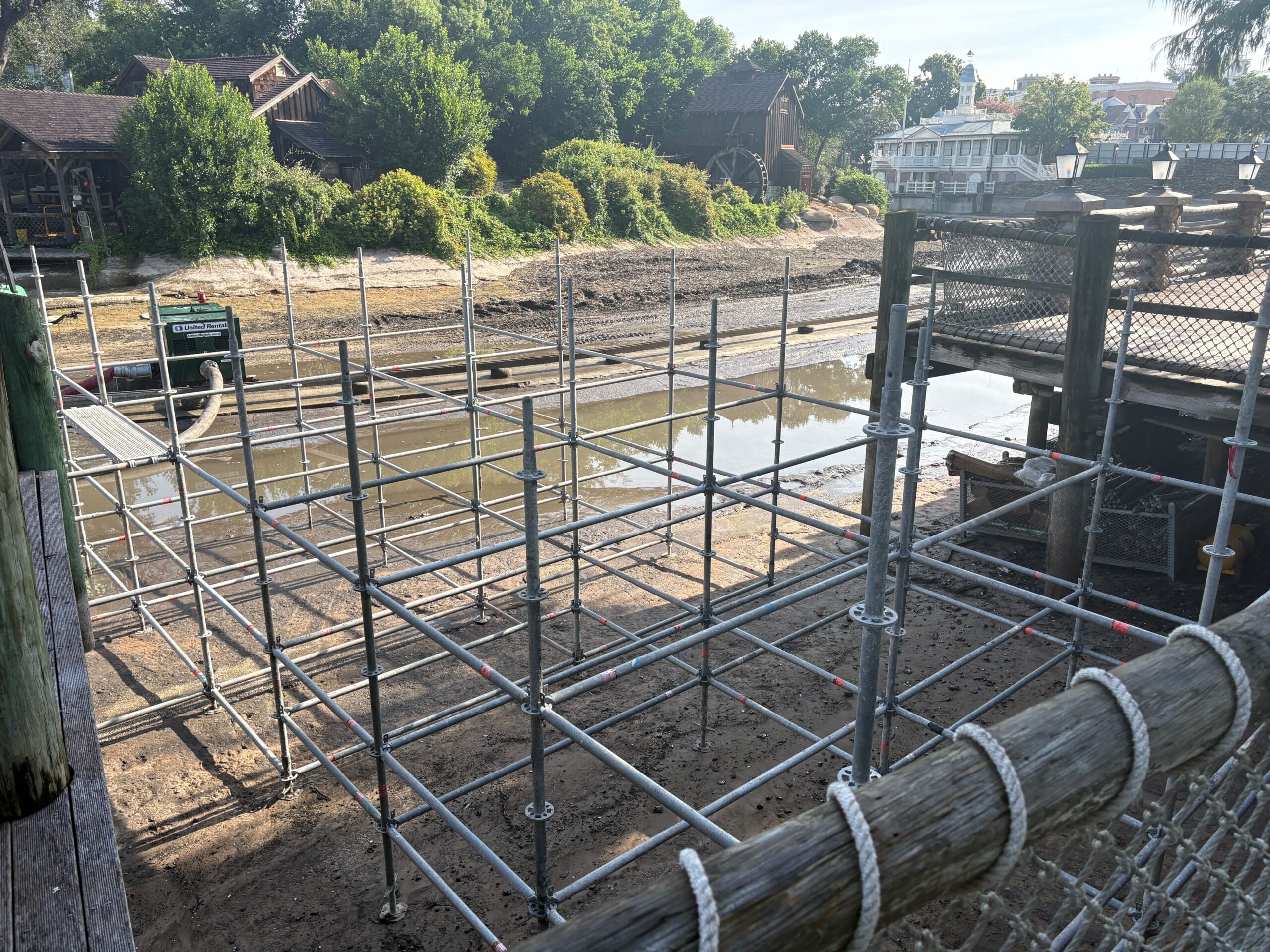 Scaffolding structure stands over a muddy area with shallow water near trees and wooden buildings.
