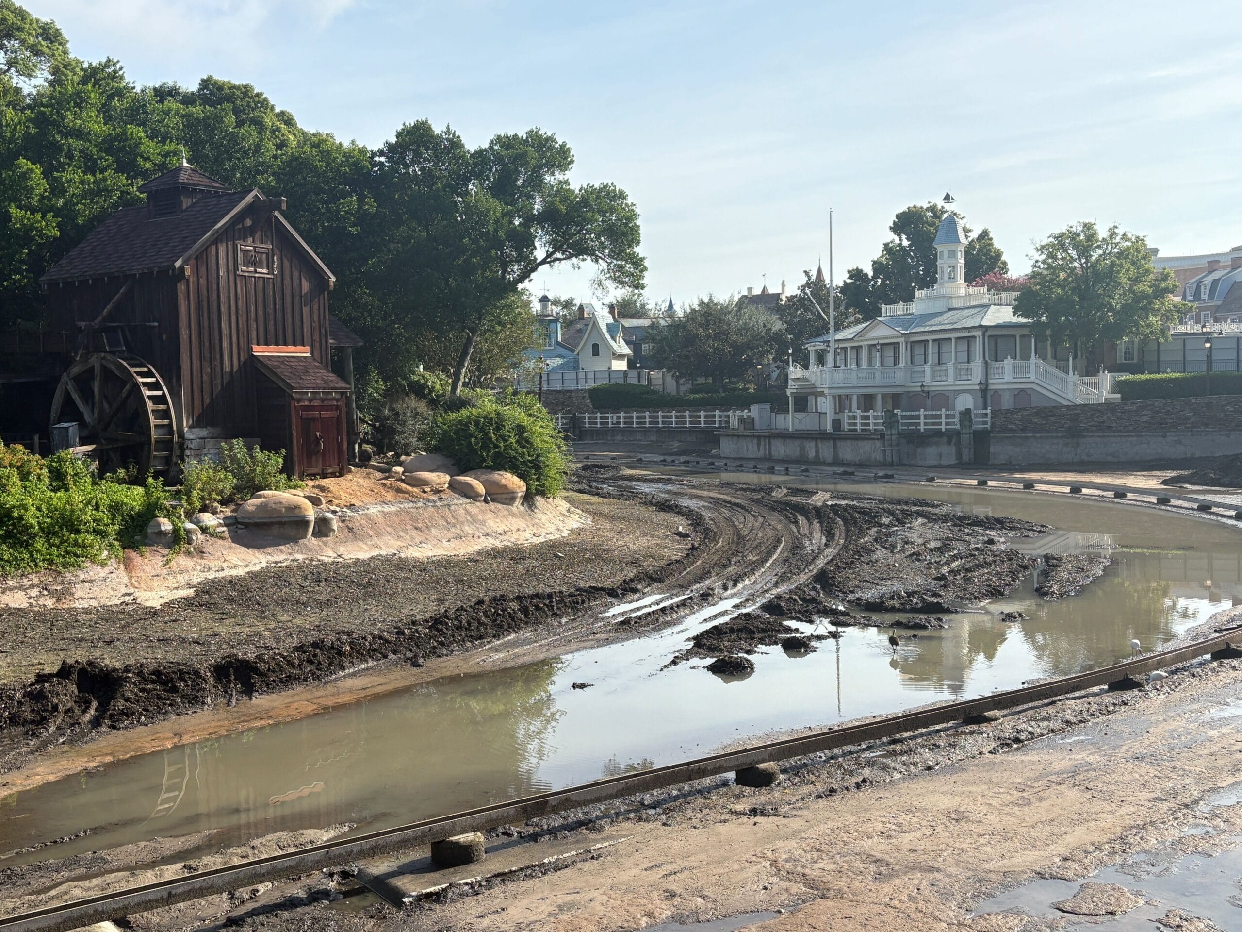 A muddy, partially drained river with a watermill and tracks, buildings, and trees in the background under a blue sky.