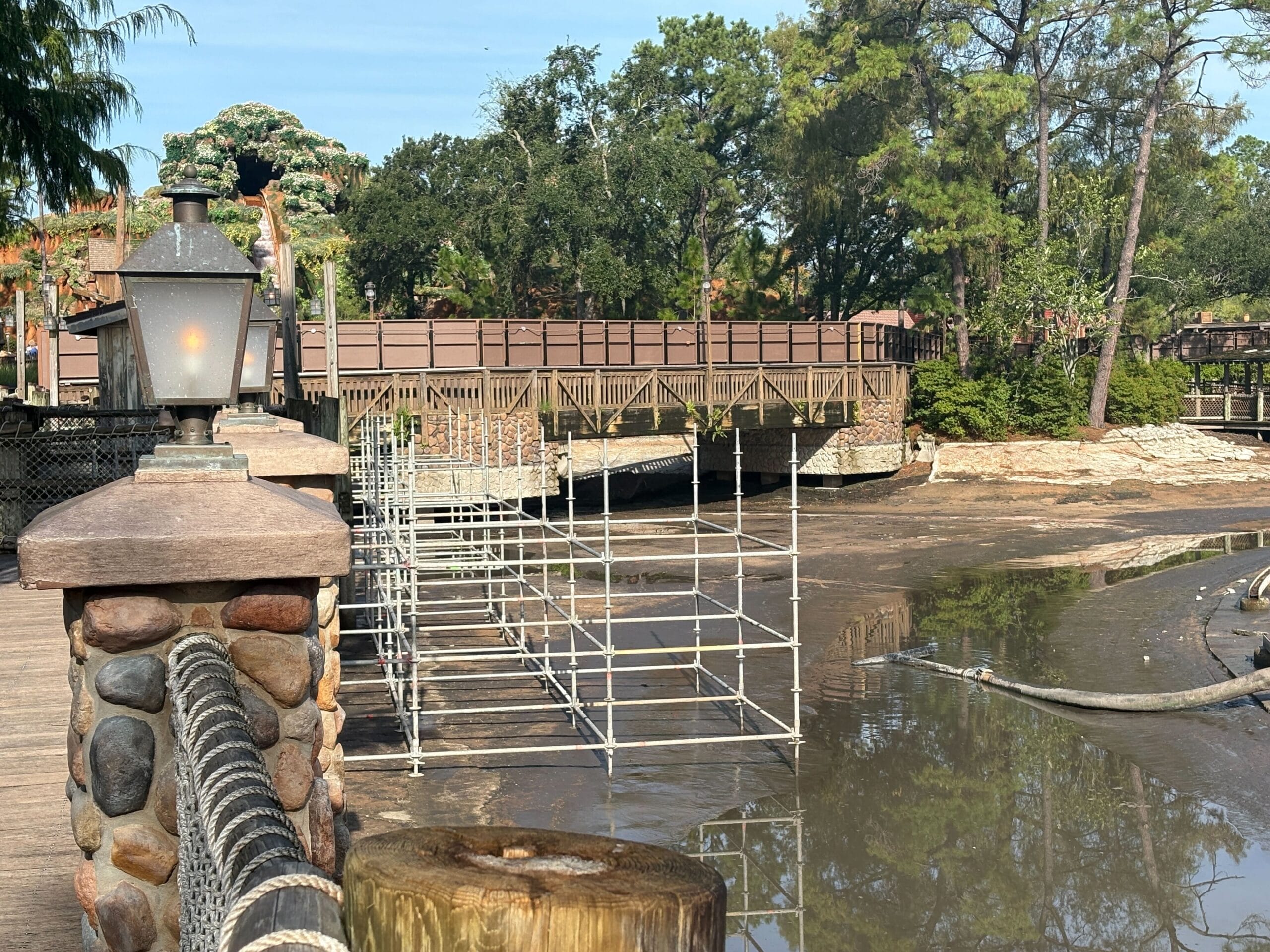 Scaffolding stands in a drained, muddy riverbed near a wooden bridge and stone lamp post, surrounded by trees.