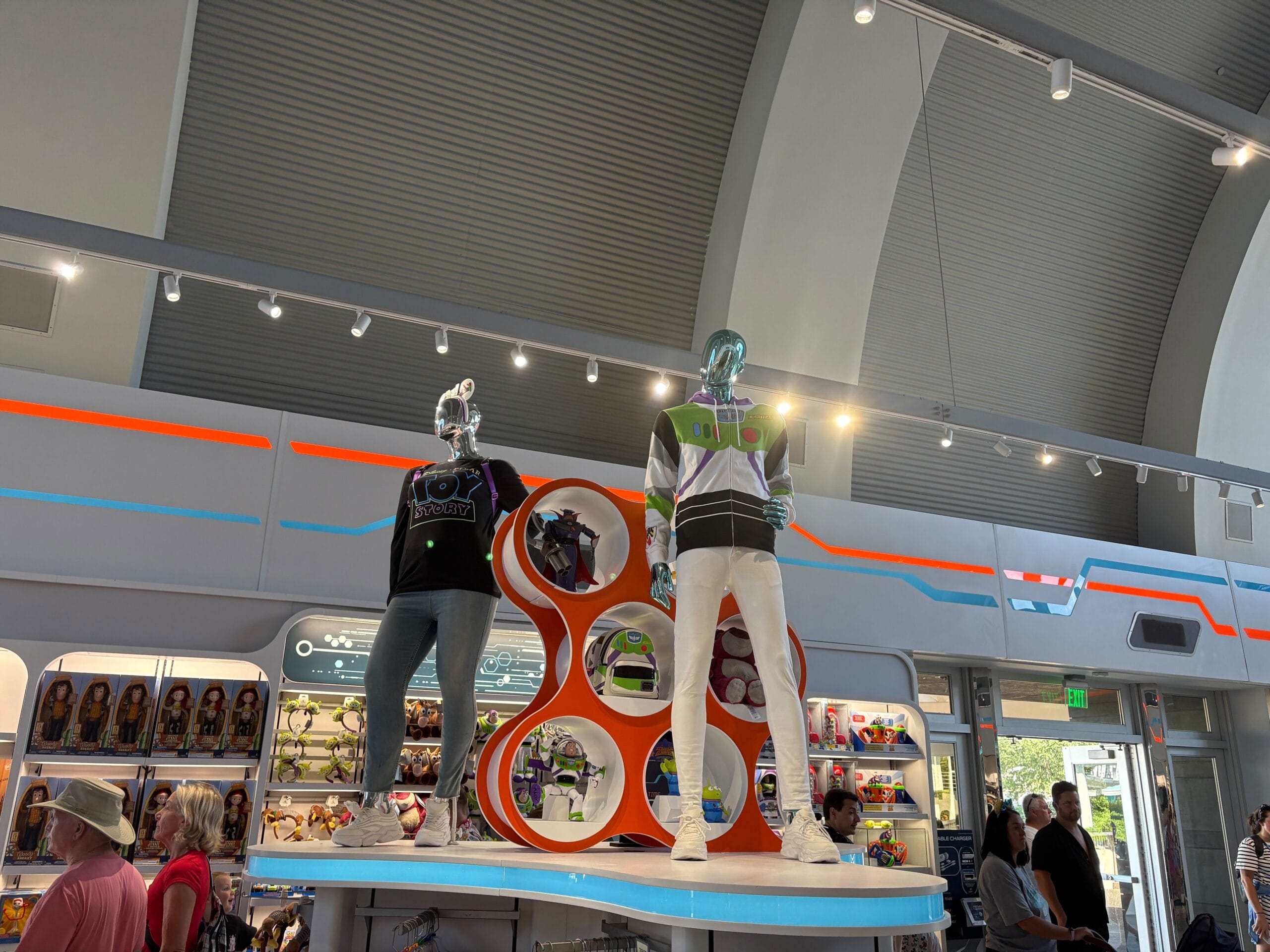 Two mannequins in futuristic clothing stand on a display in a toy store, with shelves of toys in the background.