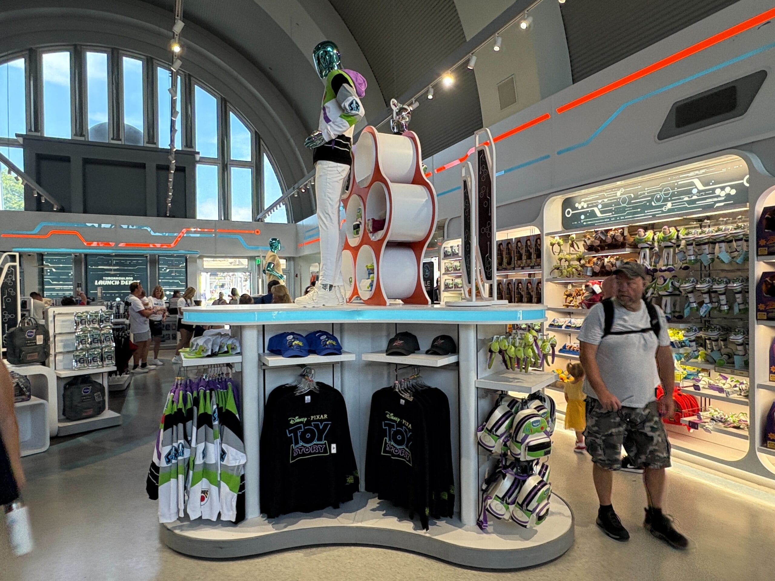 Buzz Lightyear merchandise display with hats, shirts, and toys inside a brightly lit store, shoppers visible nearby.