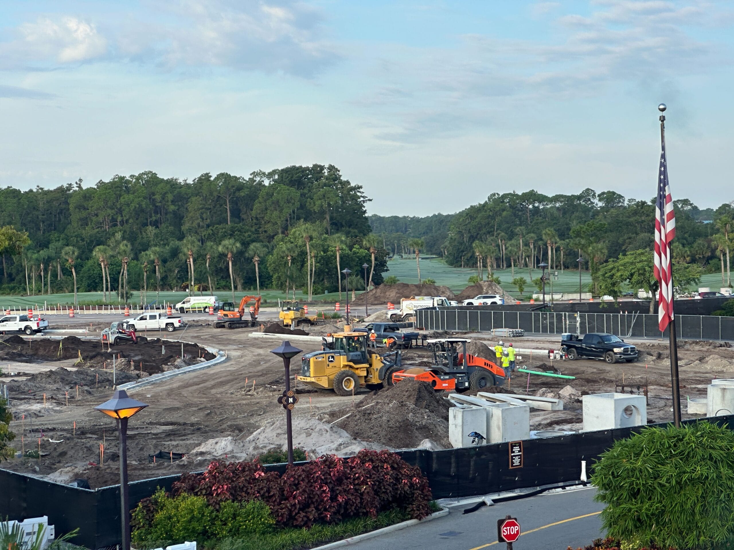 Construction entrance with heavy machinery, trucks, and workers; American flag and trees in the background.