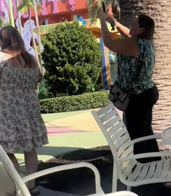 Two women stand outdoors at Pop Century, one taking a photo with her phone.