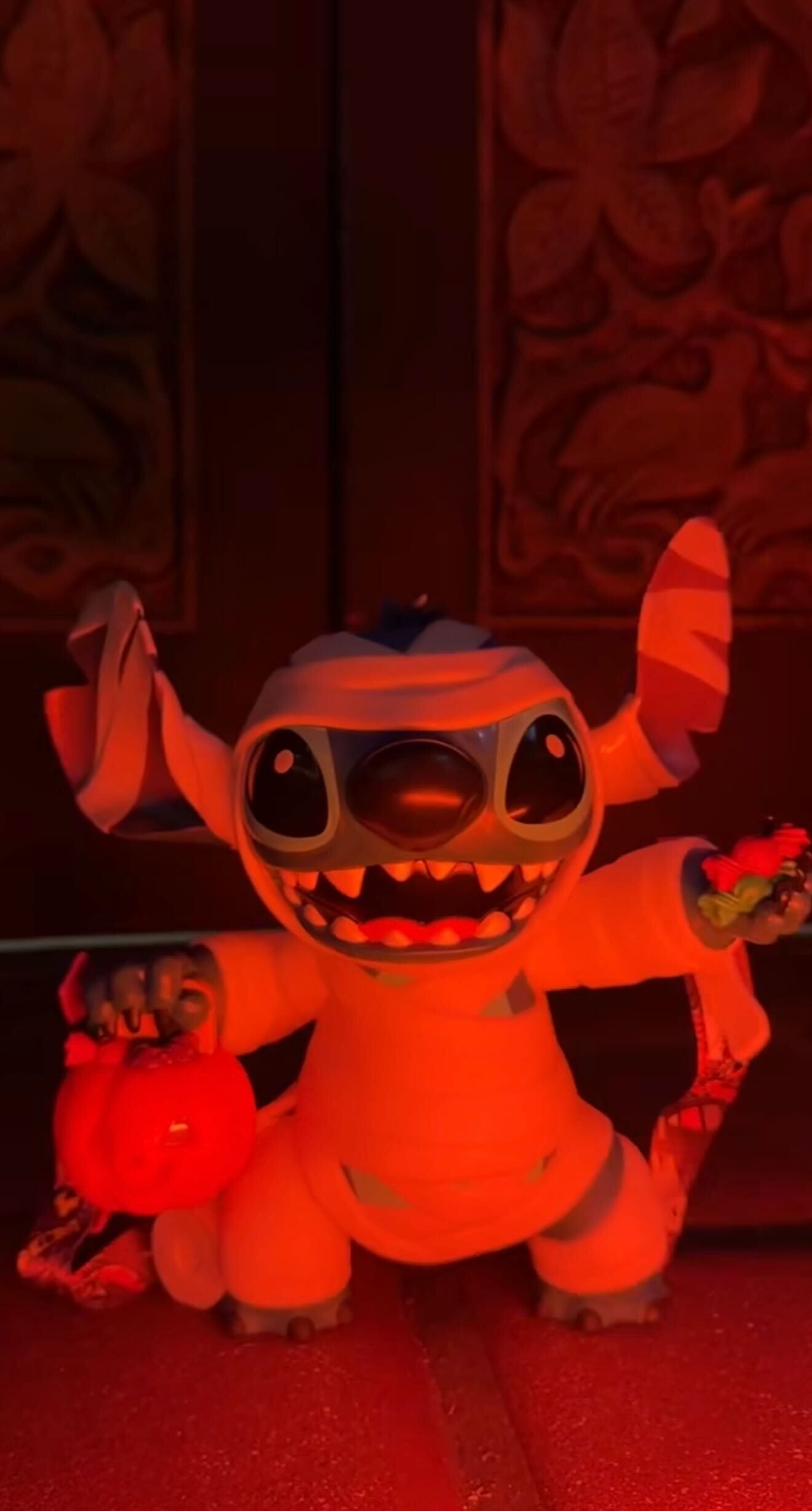 A Stitch toy dressed as a mummy holds a pumpkin bucket and candy, lit with an orange glow, in front of a carved door.