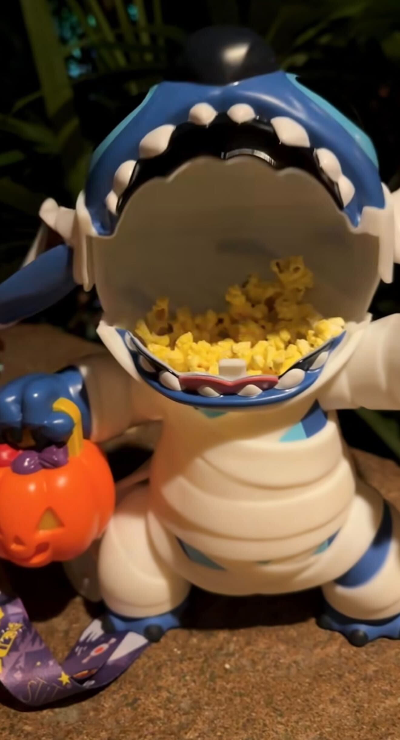 A blue and white shark toy with an open mouth holds popcorn inside and a pumpkin basket in one hand.