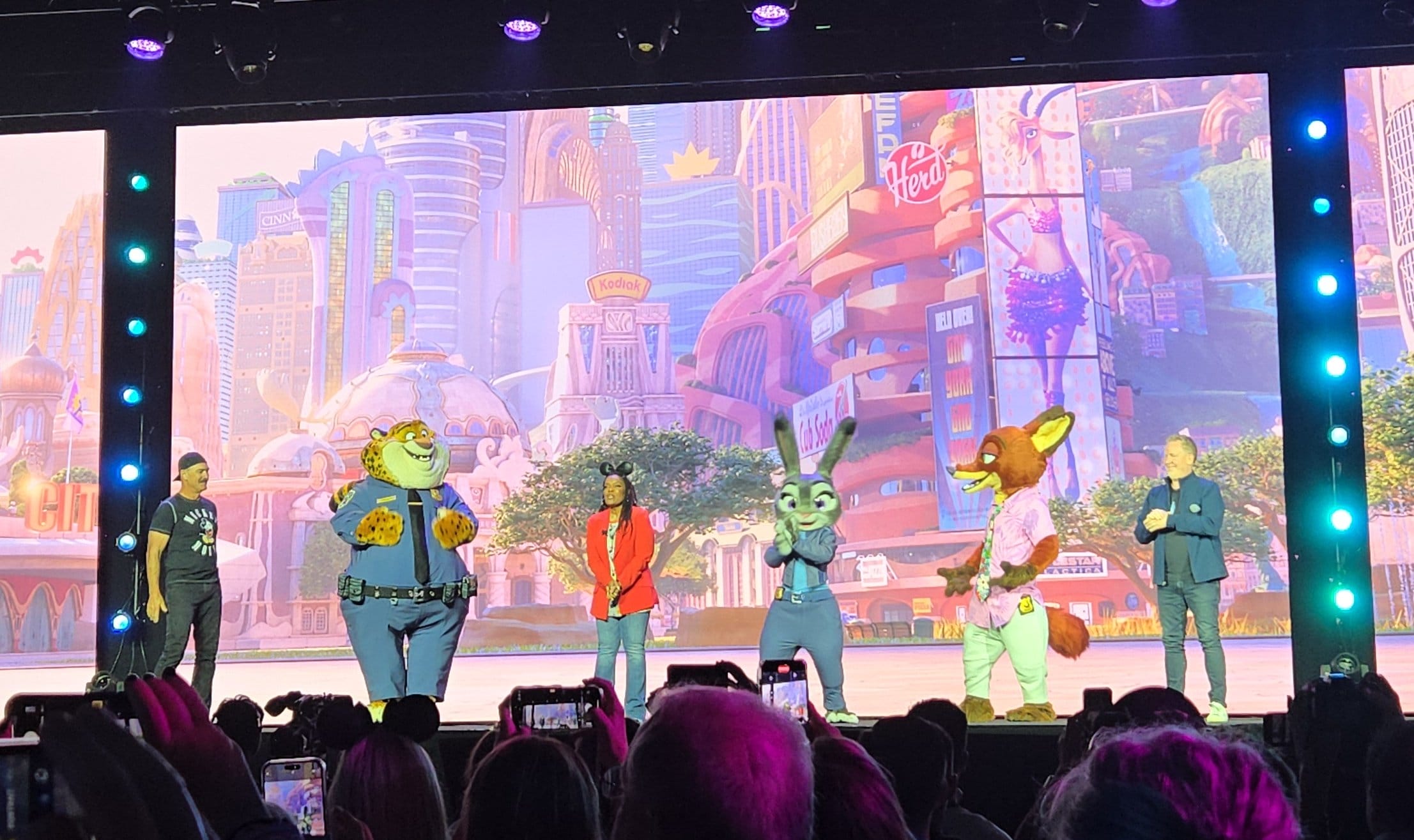People on stage with Zootopia 2's Mayor Winddancer, set against a colorful city backdrop, in front of an audience.