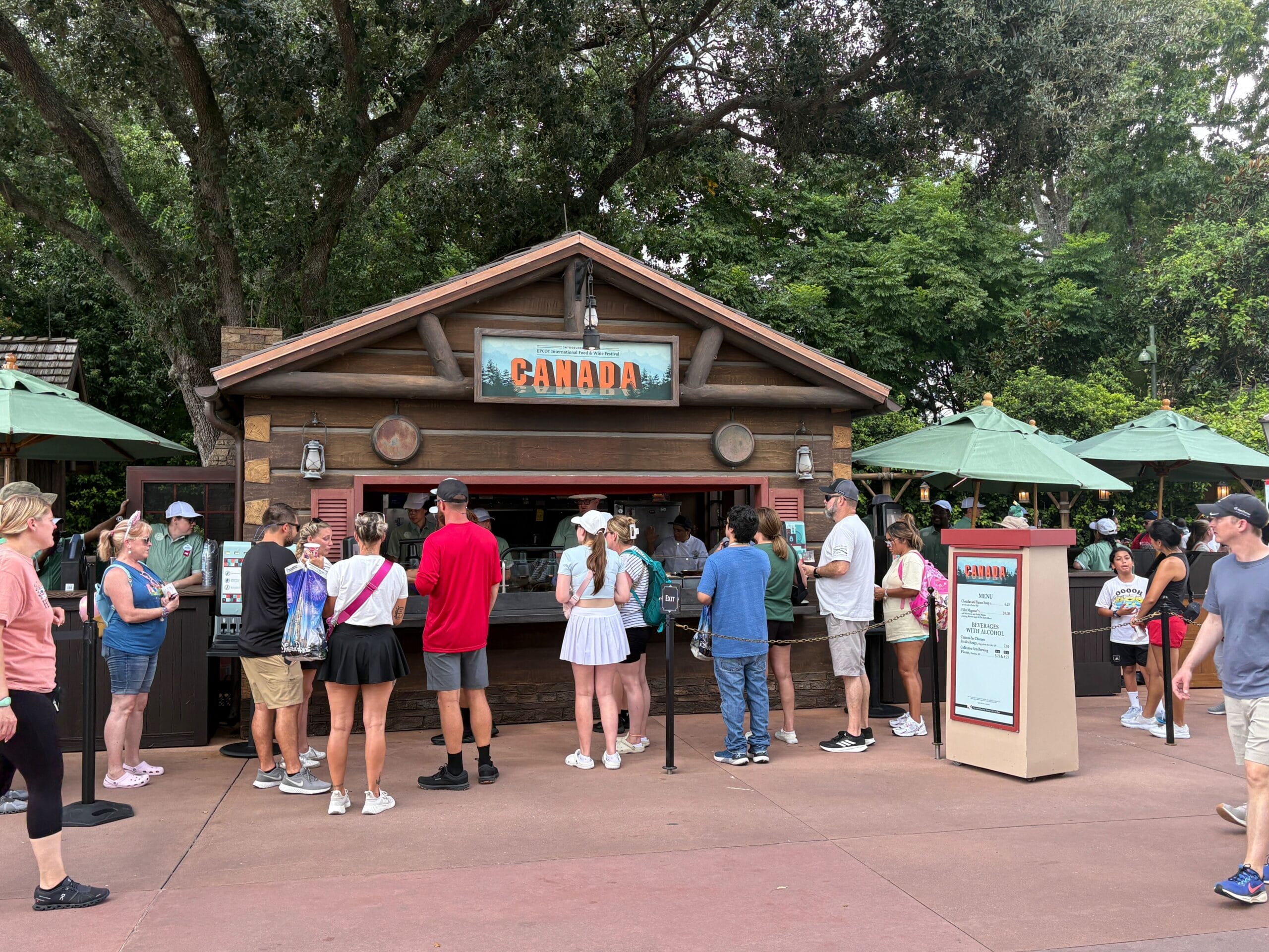 People wait in line at a Canada food stand outdoors, surrounded by trees and green umbrellas.