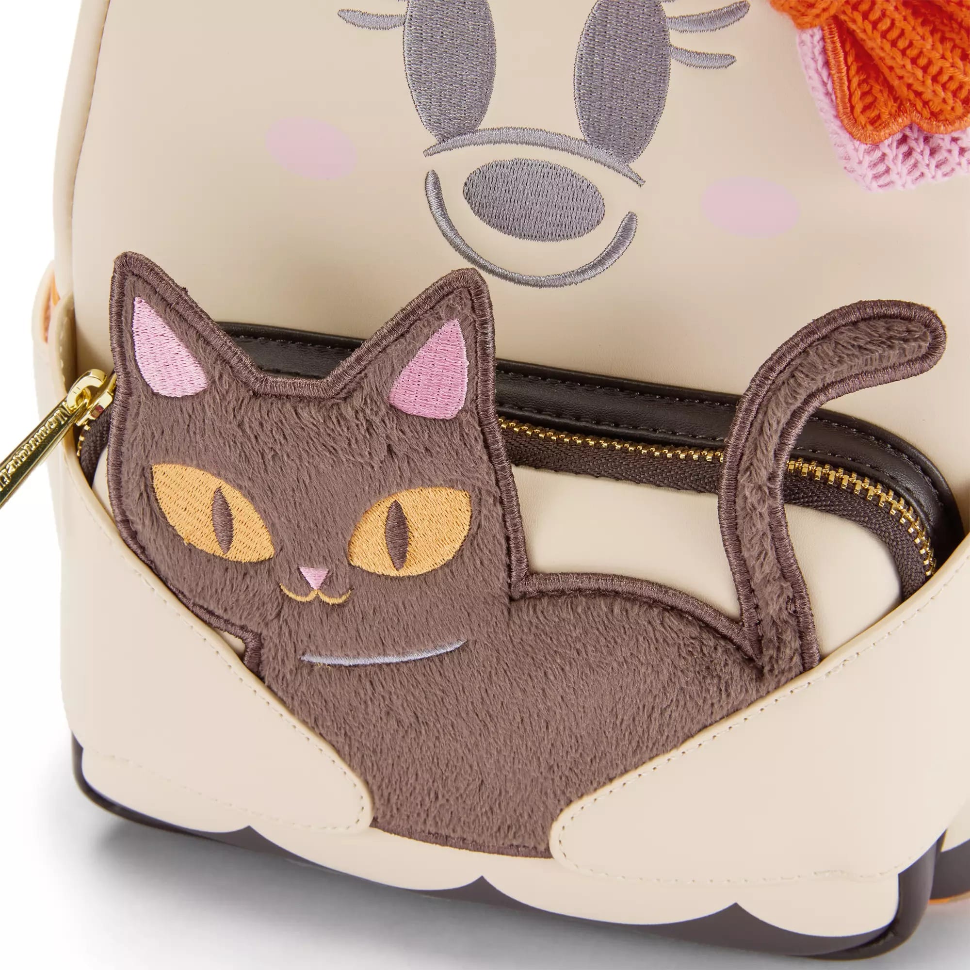 A beige bag with a stitched cartoon cat design in brown, pink, and yellow on an exterior pocket.