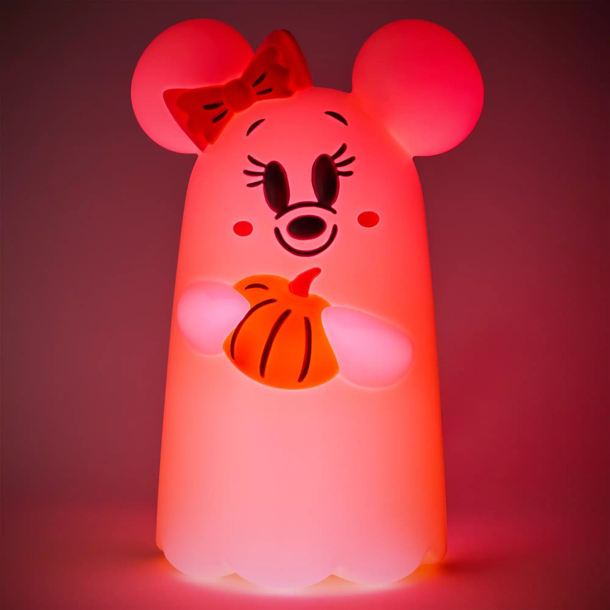 A glowing pink ghost with mouse ears, a bow, and holding an orange pumpkin, lit up in the dark.