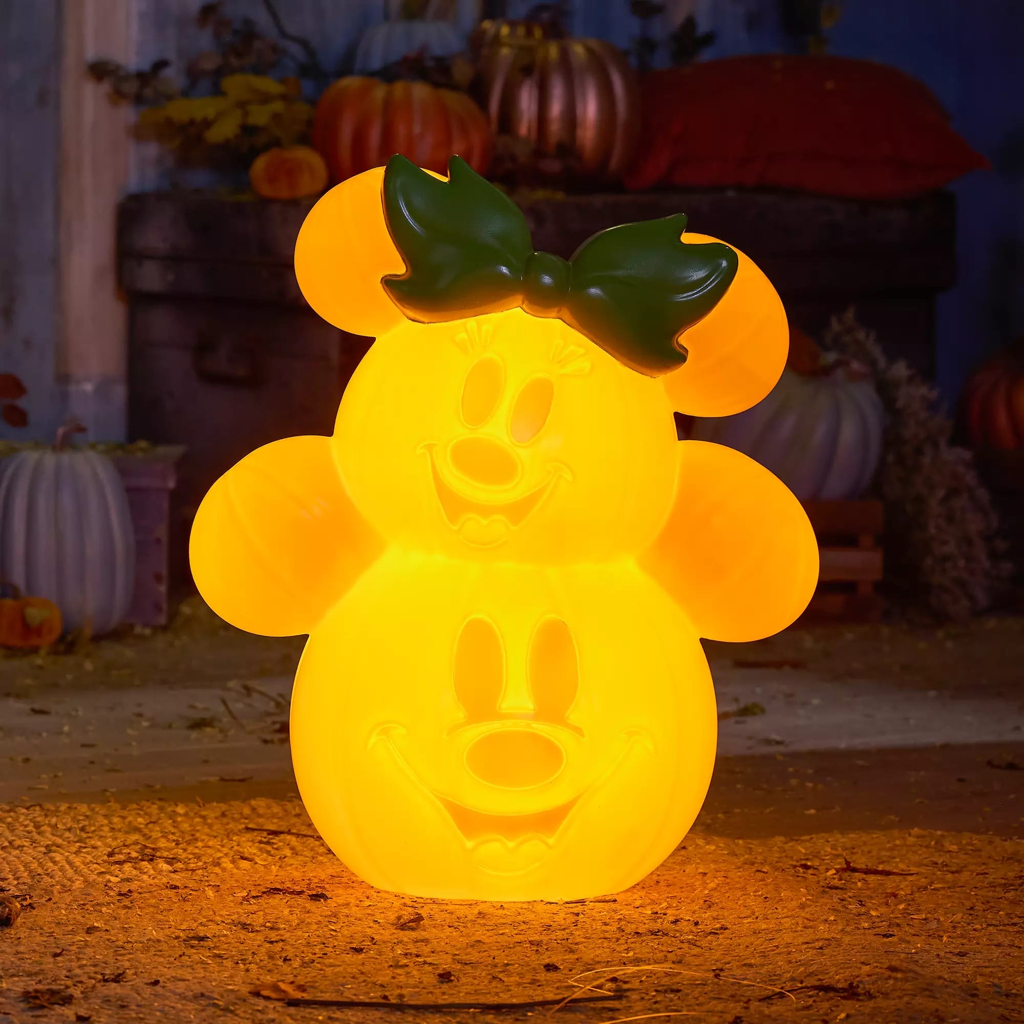 Glowing Minnie Mouse-shaped pumpkin decoration with a green bow, lit up on a rustic autumn-themed background.