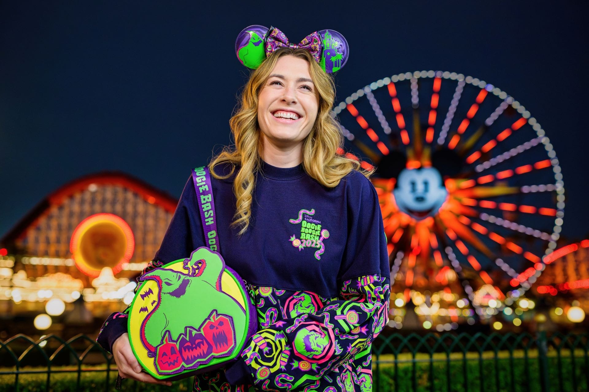 Smiling woman in colorful Halloween attire at night, with a Mickey Mouse Ferris wheel lit up in the background.