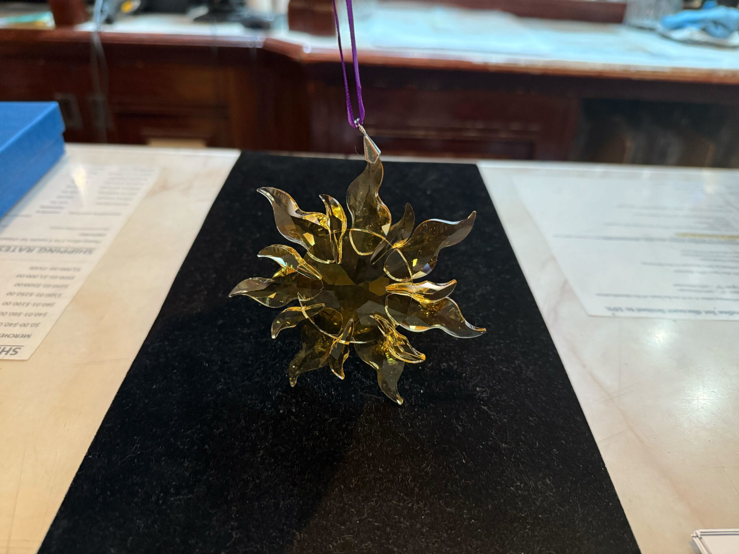 A yellow sun-shaped glass ornament with wavy rays hangs by a purple string over a black surface.