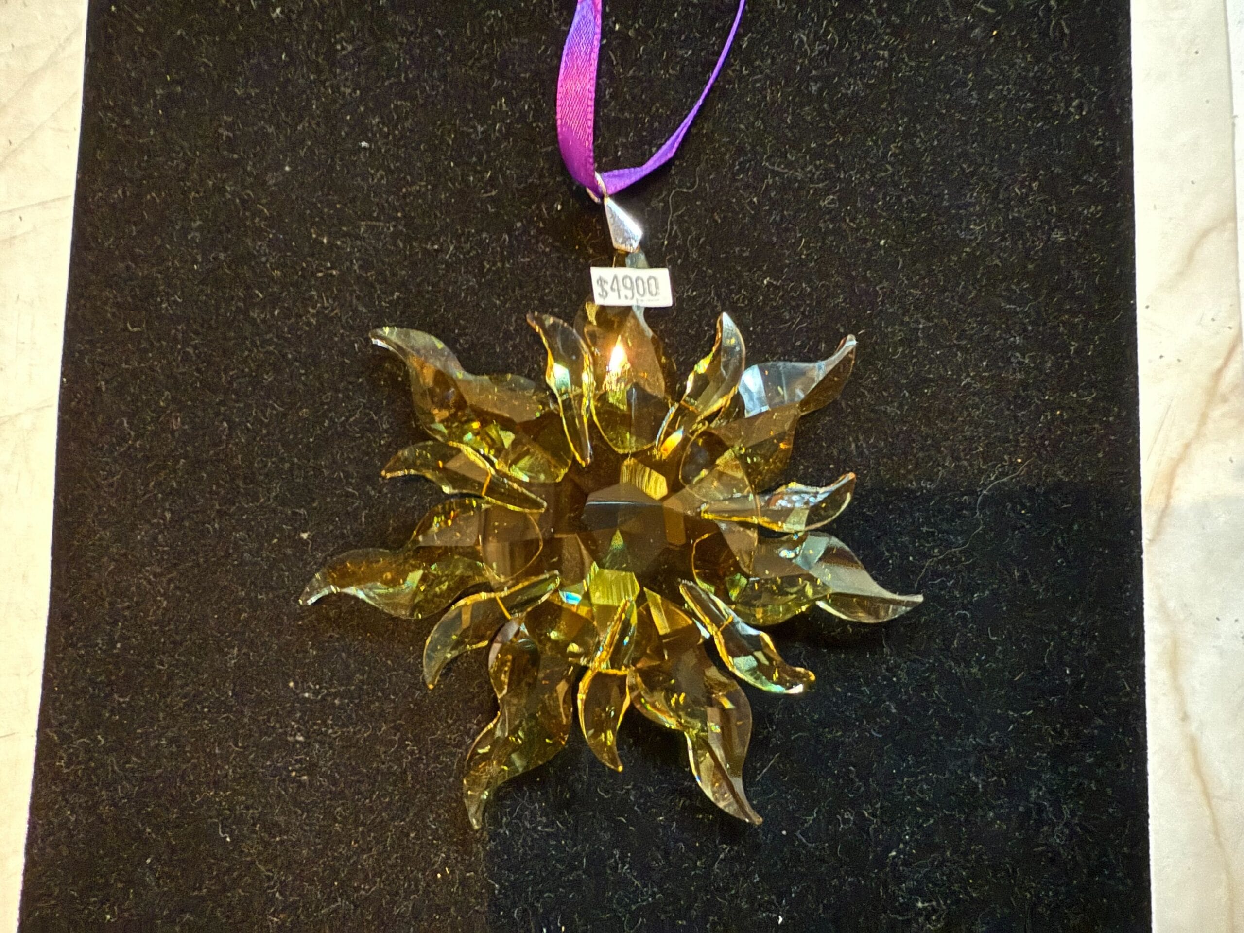 A yellow glass sun-shaped ornament with a $49.00 tag and a purple ribbon, displayed on black fabric.