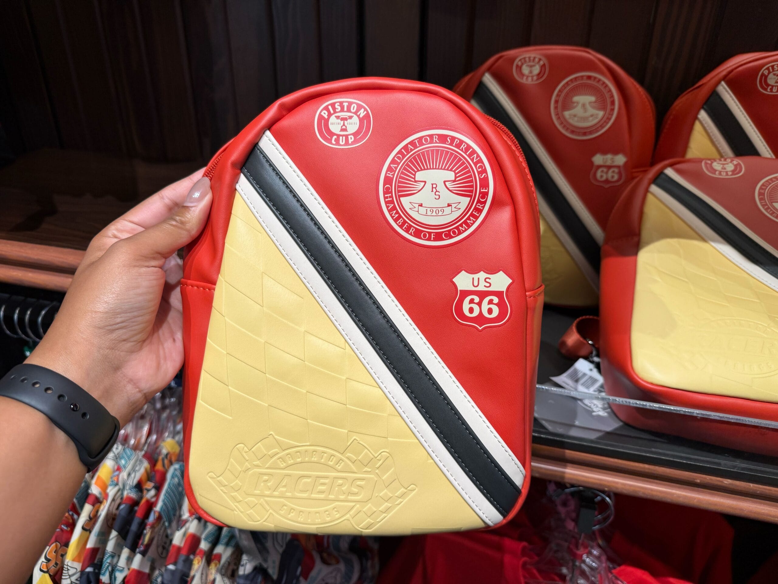 Hand holding a small red, yellow, and black backpack with Route 66, piston cup, and RACERS logo designs.
