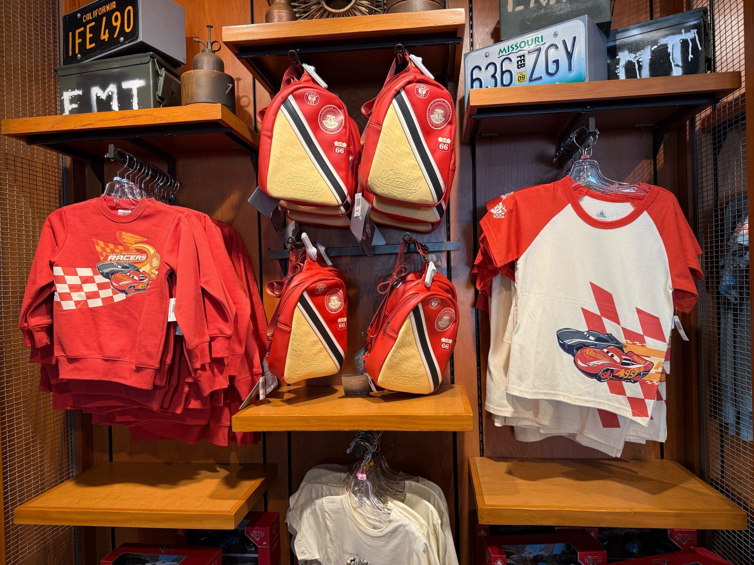 Red and white Cars-themed shirts and crossbody bags are displayed on wooden shelves in a store.