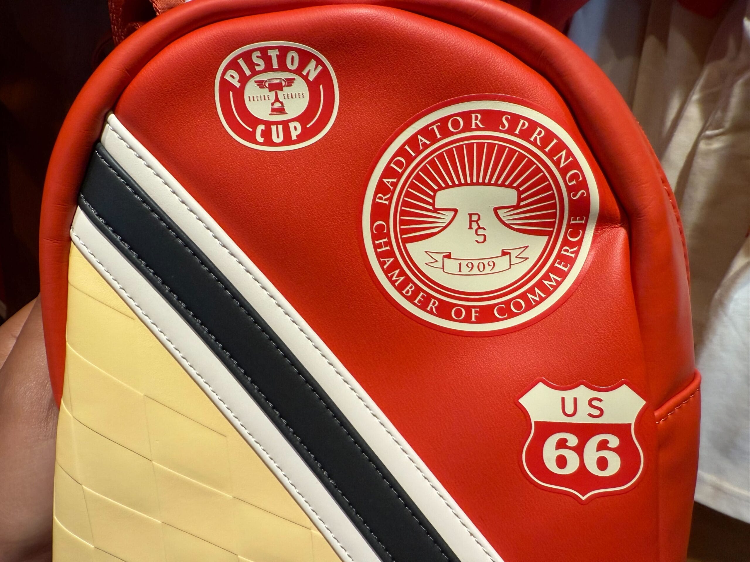 Red backpack with Piston Cup, Radiator Springs, and US Route 66 logos, featuring yellow, black, and white diagonal stripes.