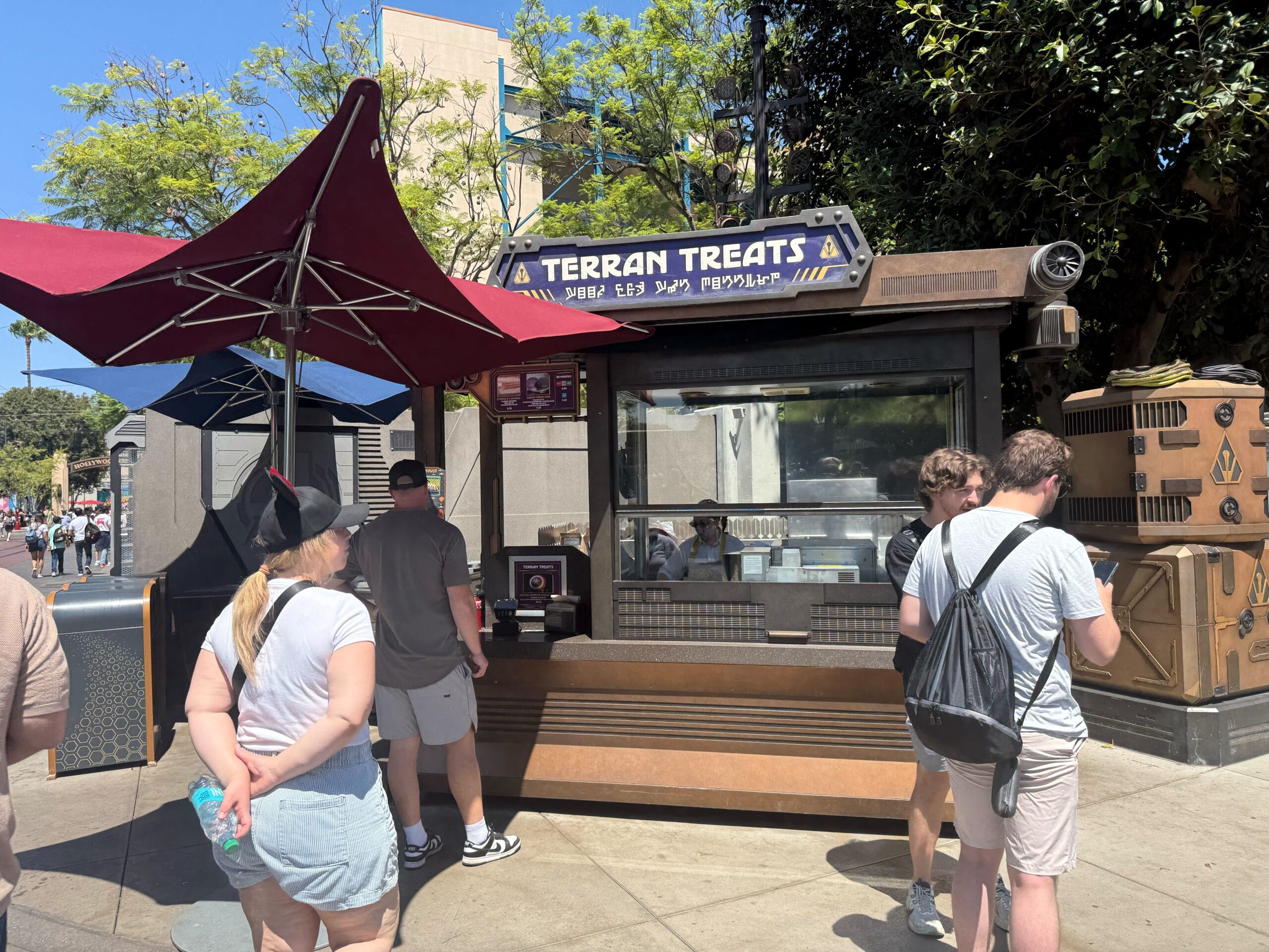 People wait in line at Disney California Adventure’s Terran Treats for a Celestial Chocolate Churro under a red umbrella.