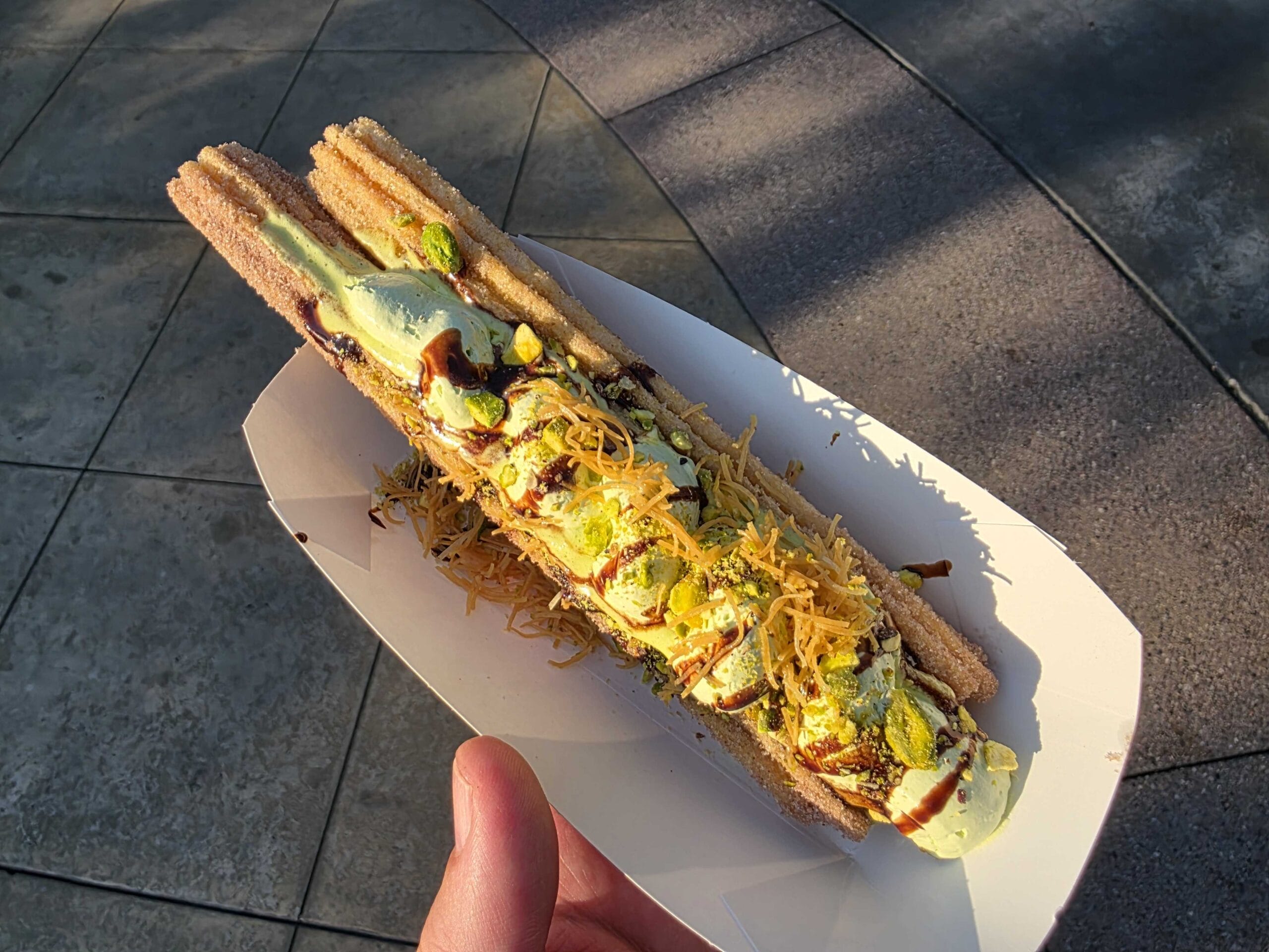 A hand holds a Celestial Chocolate Churro topped with cream, cheese, sauce, and pistachios at Disney California Adventure.