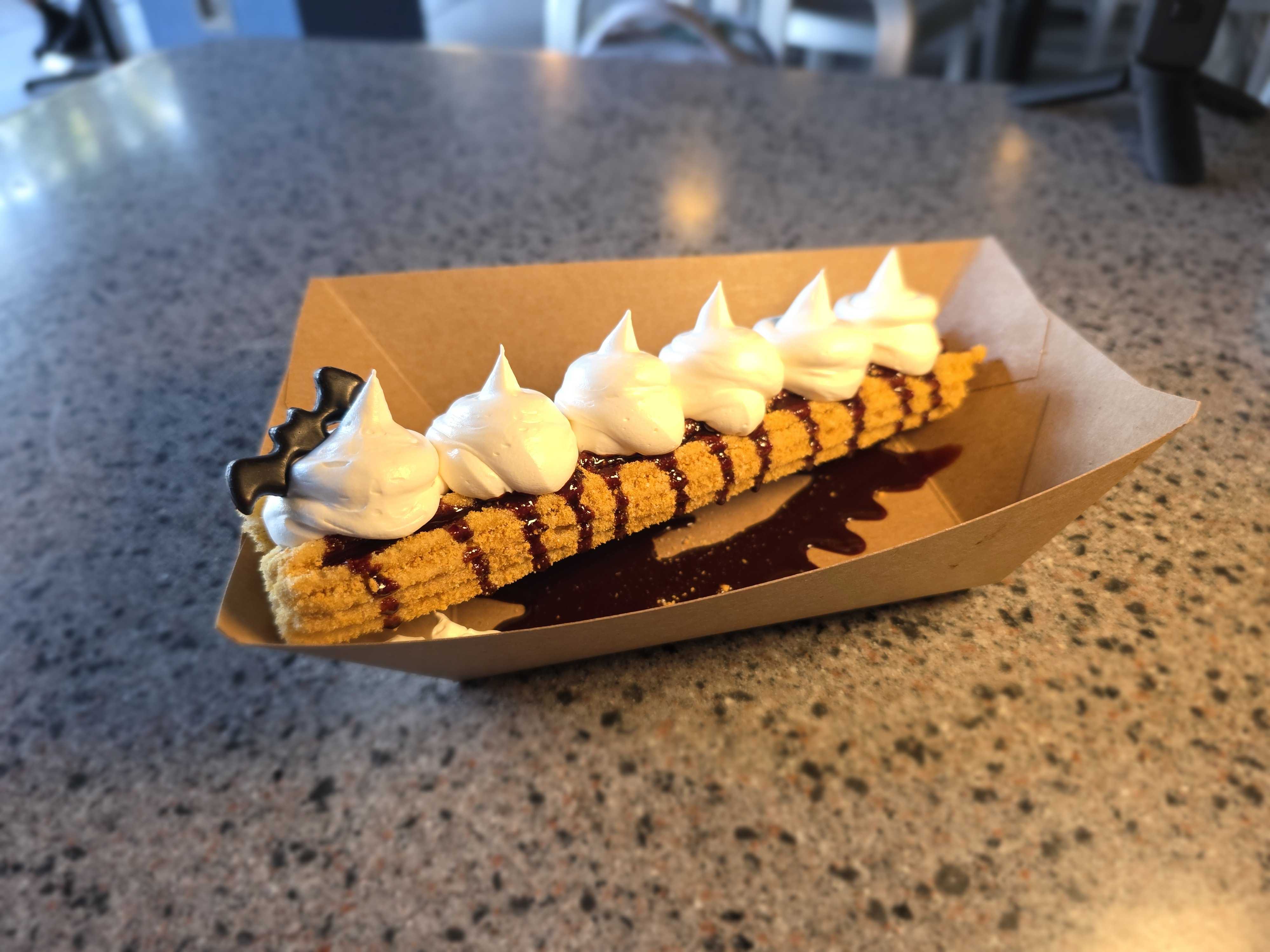 A DL Halloween churro topped with chocolate sauce and whipped cream in a brown paper tray on a speckled counter.