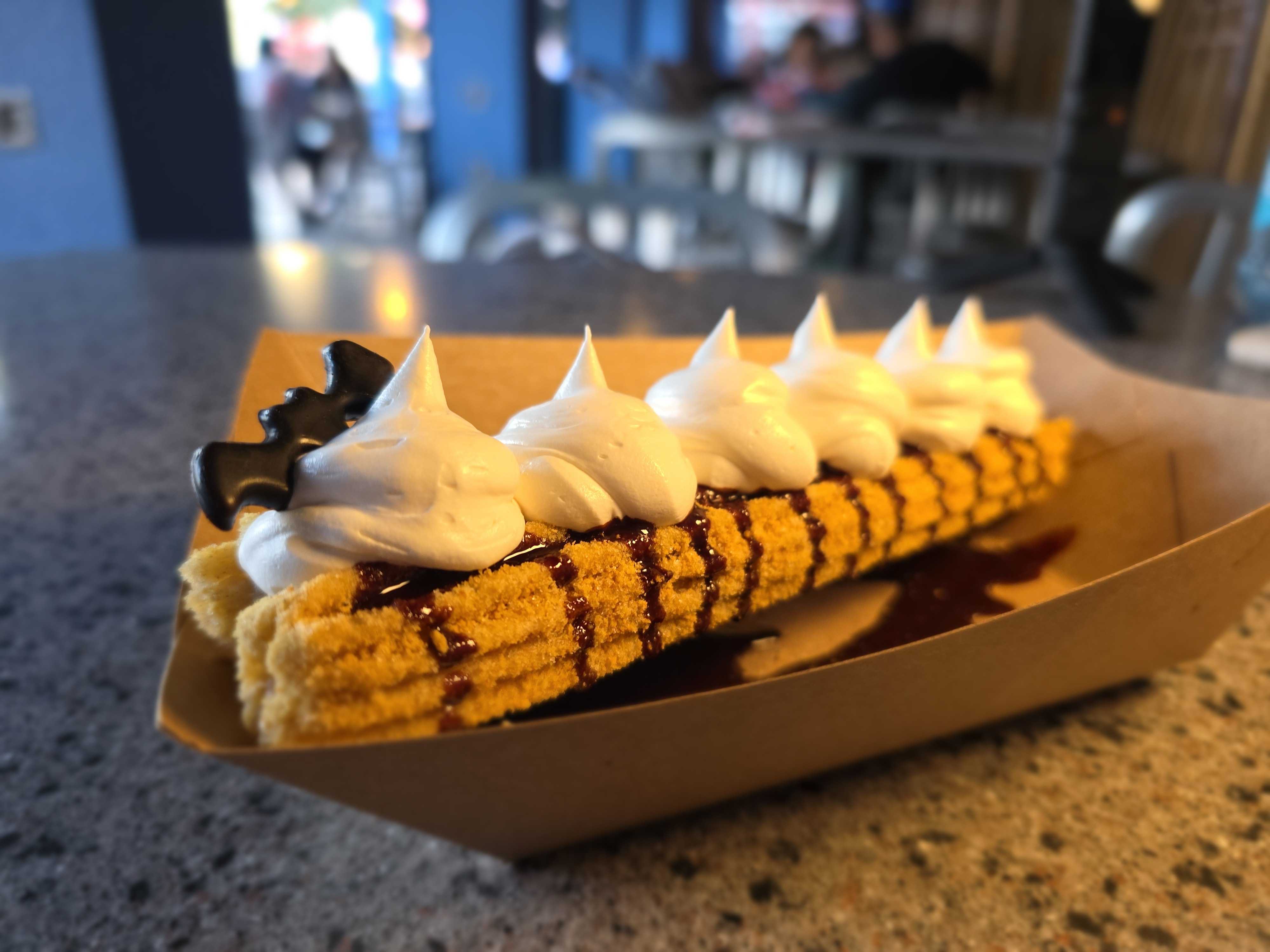 A DL Halloween churro topped with whipped cream, chocolate drizzle, and a bat-shaped chocolate piece in a brown tray.