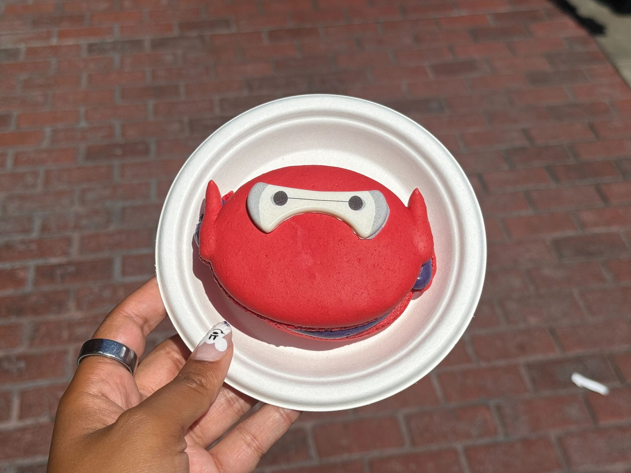 A hand holds a red Ube Baymax Macaron at Disney California Adventure, set on a white plate with brick ground behind.