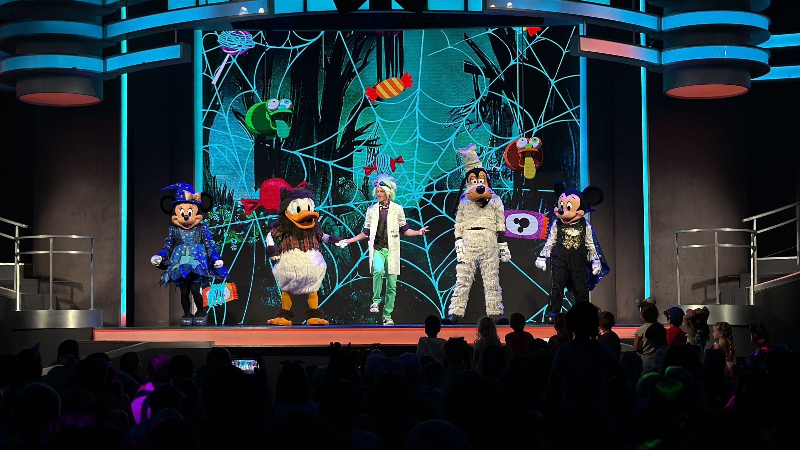 Five Disney characters in Halloween costumes perform on stage at a Disneyland Halloween event with a spiderweb backdrop.