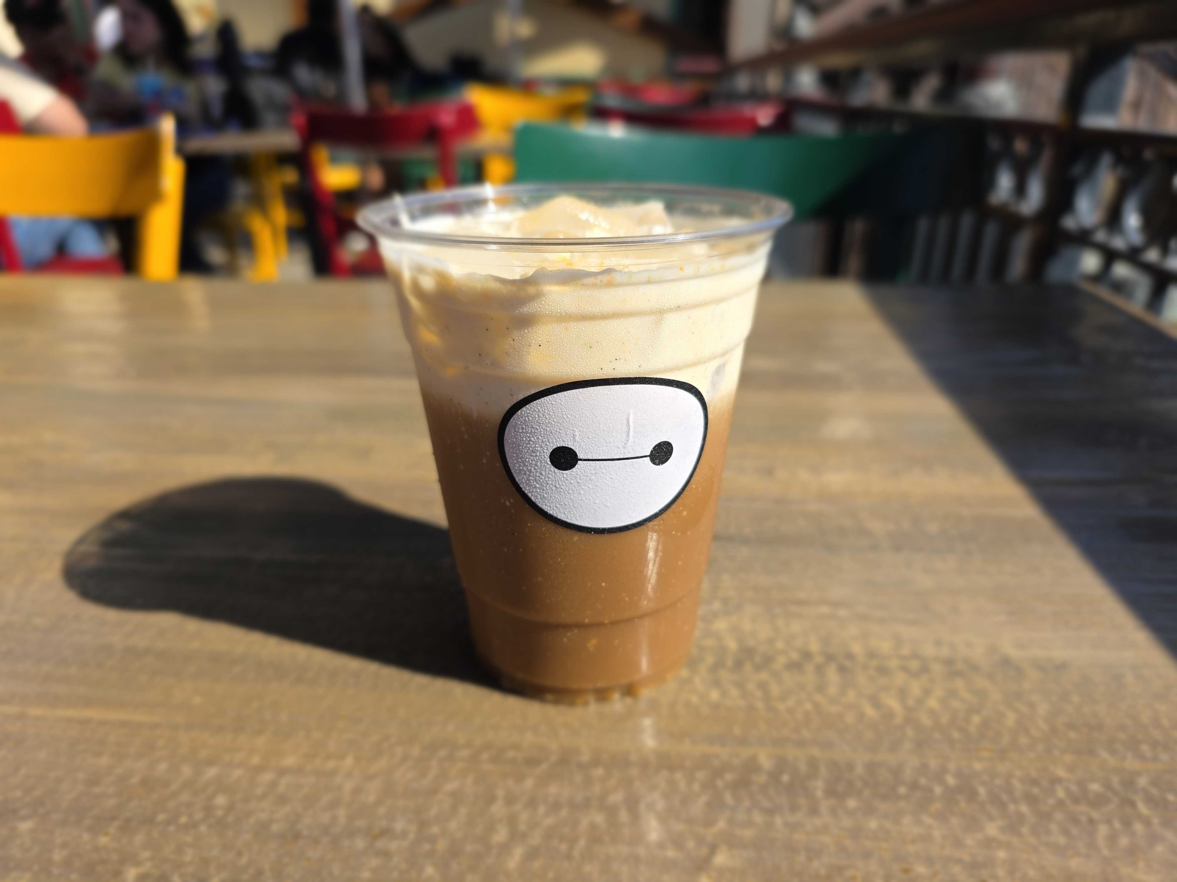 A plastic cup of iced coffee with cream from Aunt Cass Cafe sits on a wooden table, featuring a cartoon face.