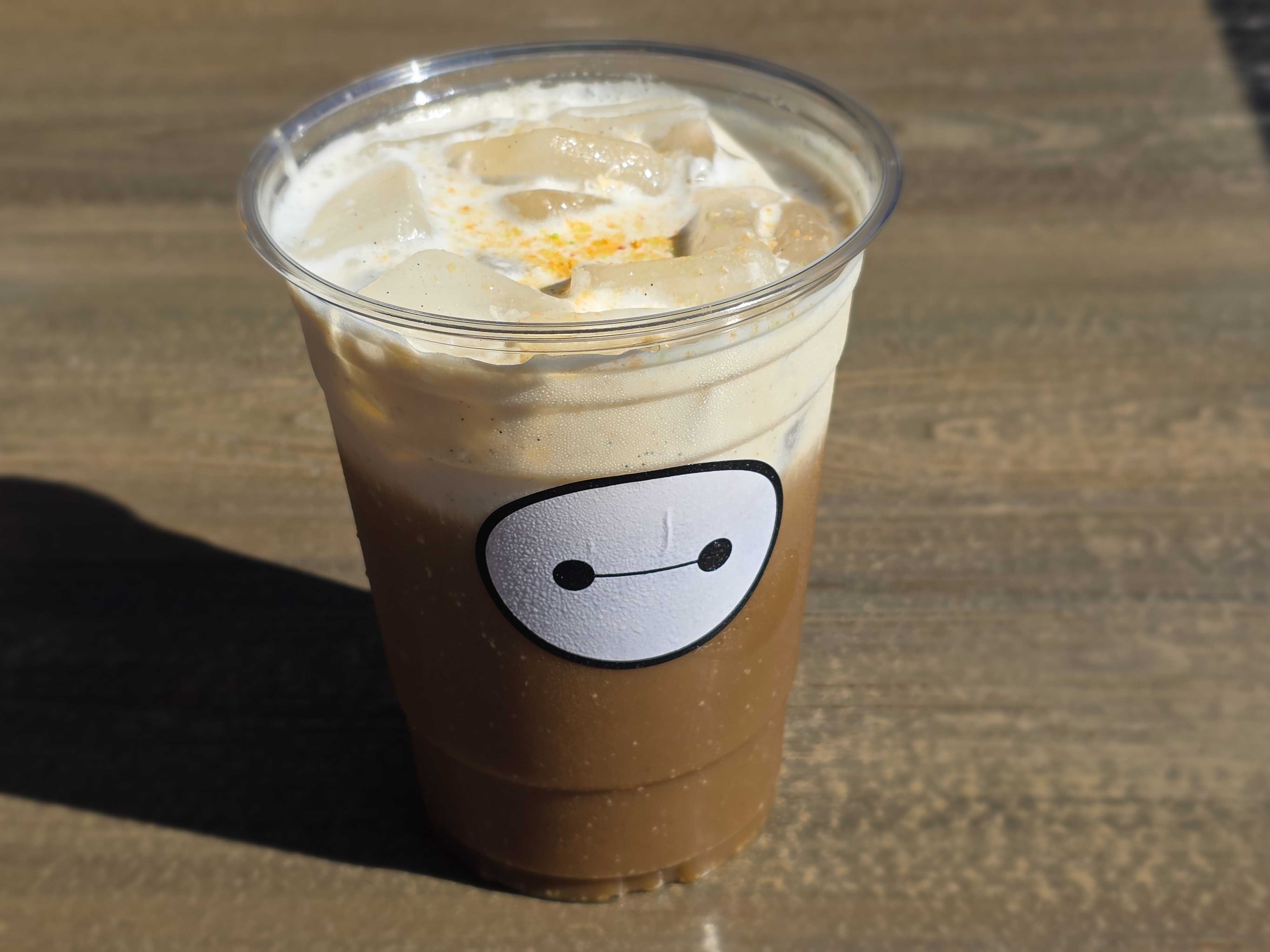 Iced coffee in a clear cup with a Baymax face sticker, served at Aunt Cass Cafe on a wooden surface.