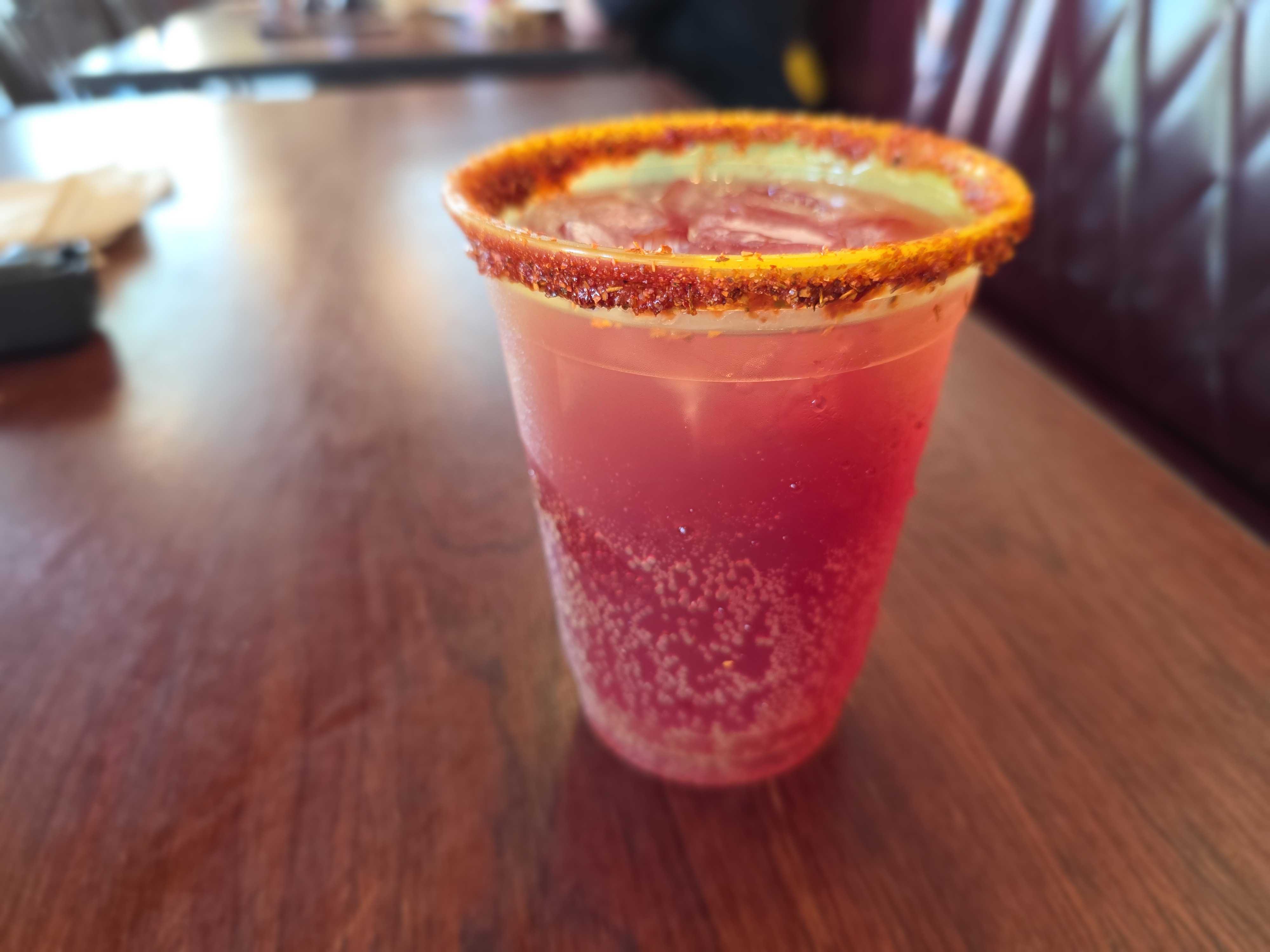 A red, fizzy Blackberry Margarita with ice in a plastic cup, rimmed with chili powder, sits on a wooden table.