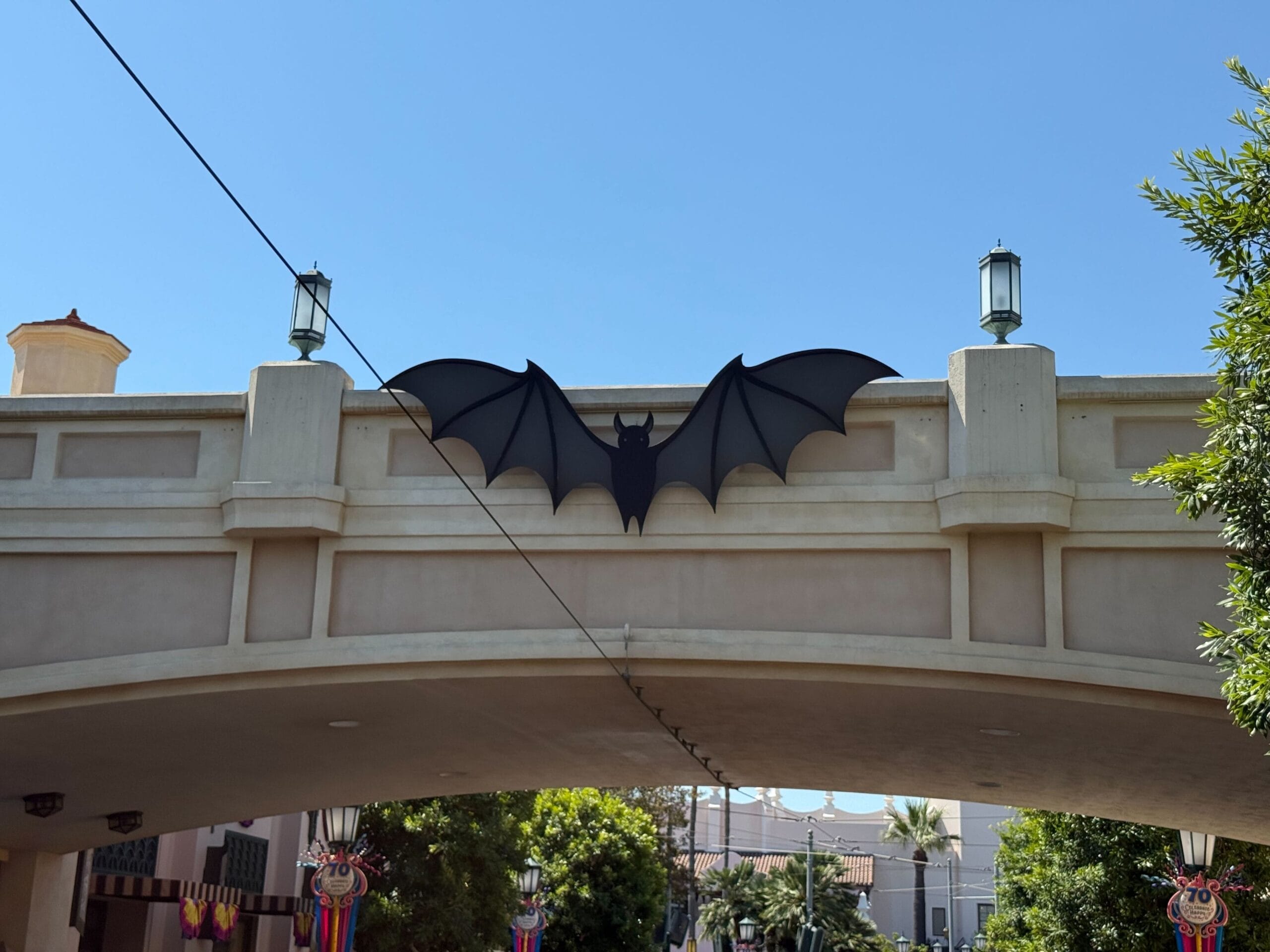 A large black bat decoration is displayed on a bridge across Buena Vista Street in Disney California Adventure, with trees and lanterns nearby.