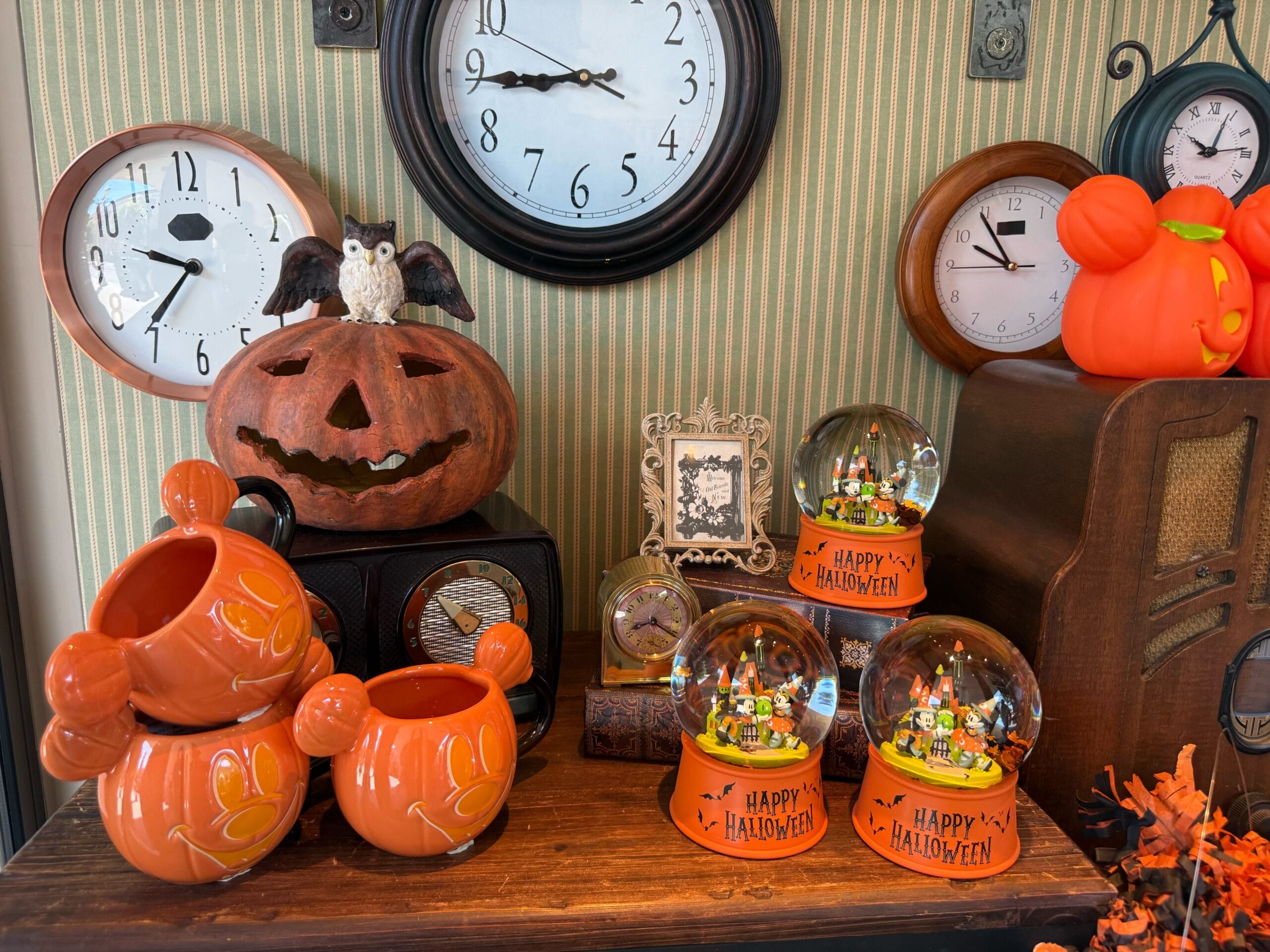 Halloween decorations with pumpkin mugs, snow globes, jack-o-lanterns, and clocks on a wooden table.