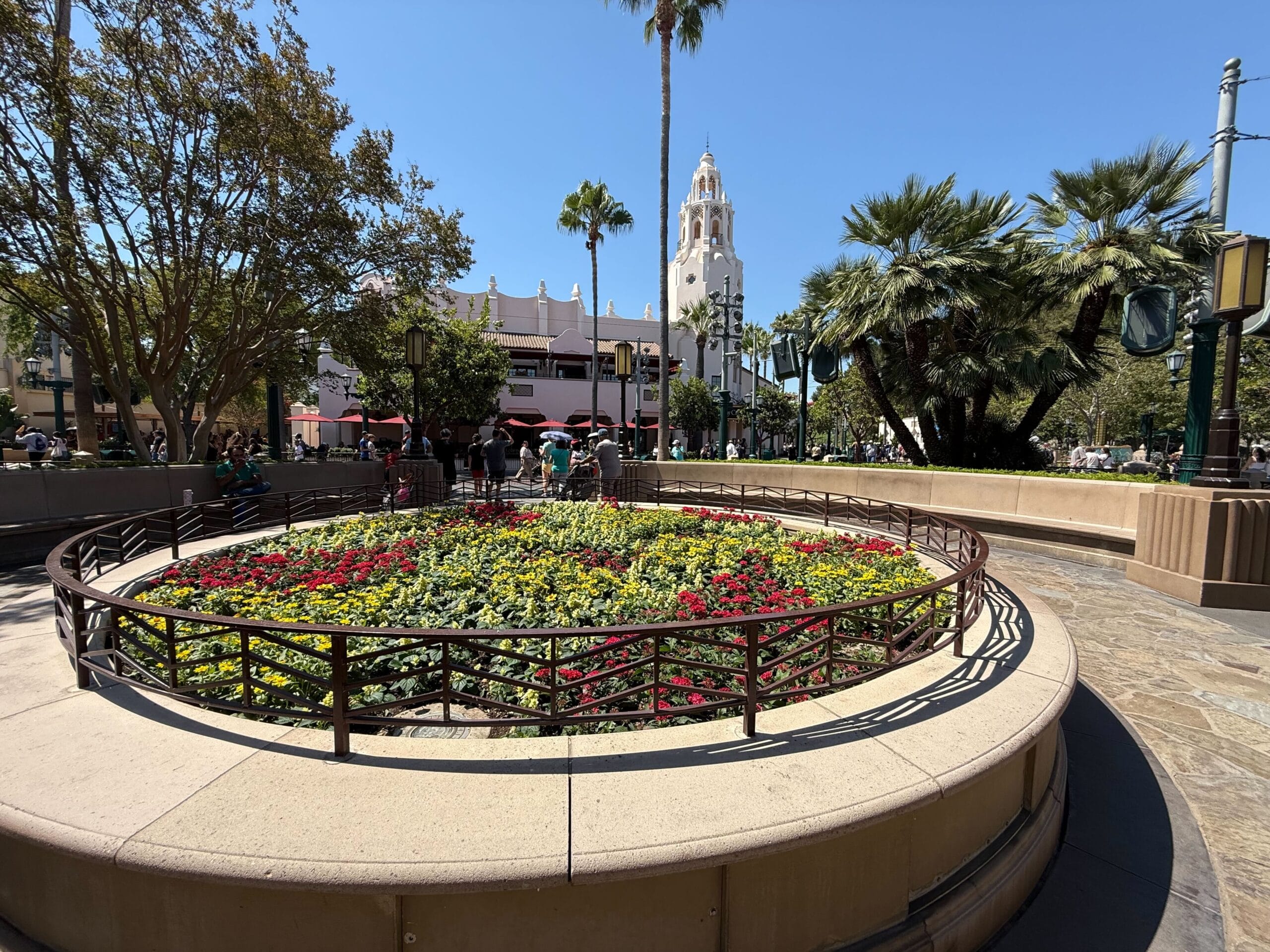 A round flower bed with colorful blooms in a plaza of Disney California Adventure, with palm trees and Carthay Circle Restaurant in the background.