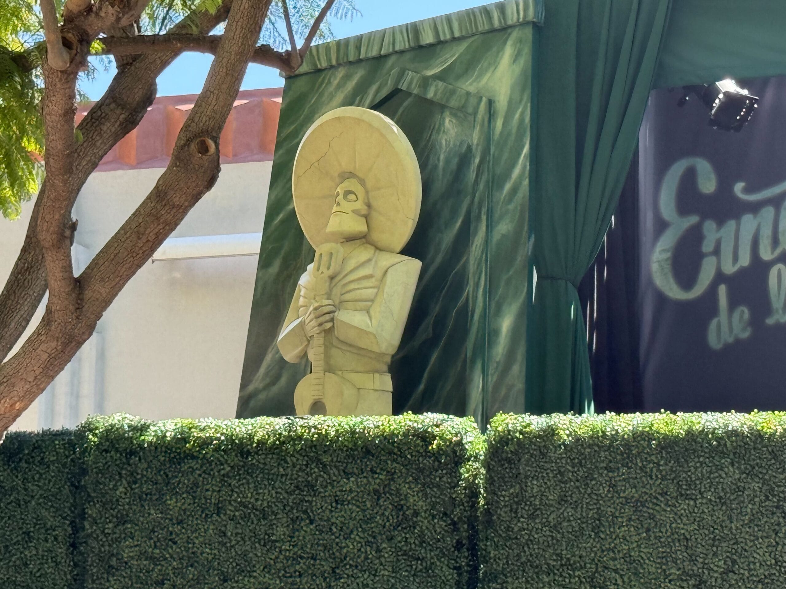 A skeleton statue of Ernesto de la Cruz with a large hat holds a guitar, standing by green curtains and a leafy hedge.