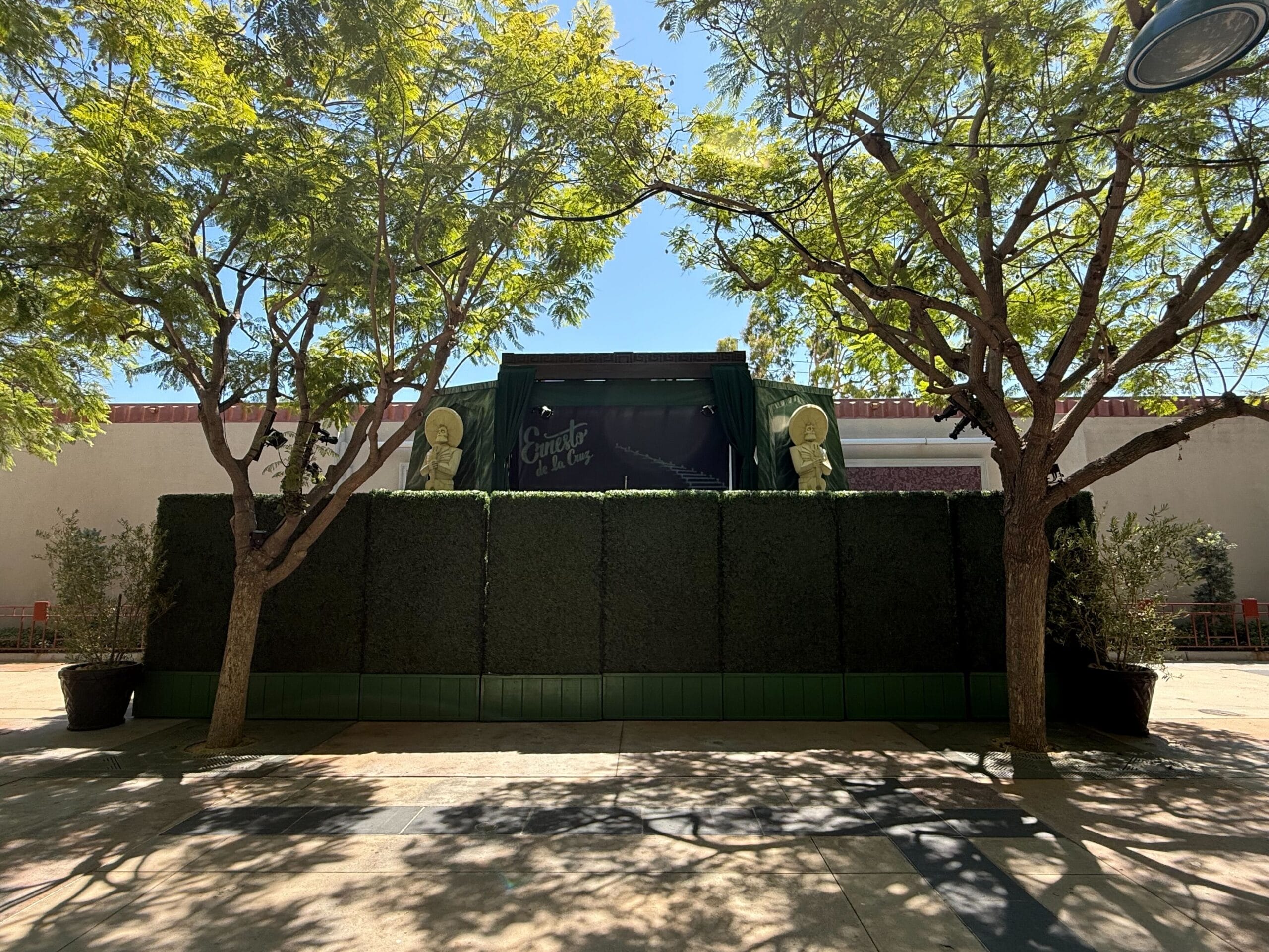 A small outdoor stage with greenery, flanked by trees, under bright sunlight in an open plaza of Disney California Adventure.