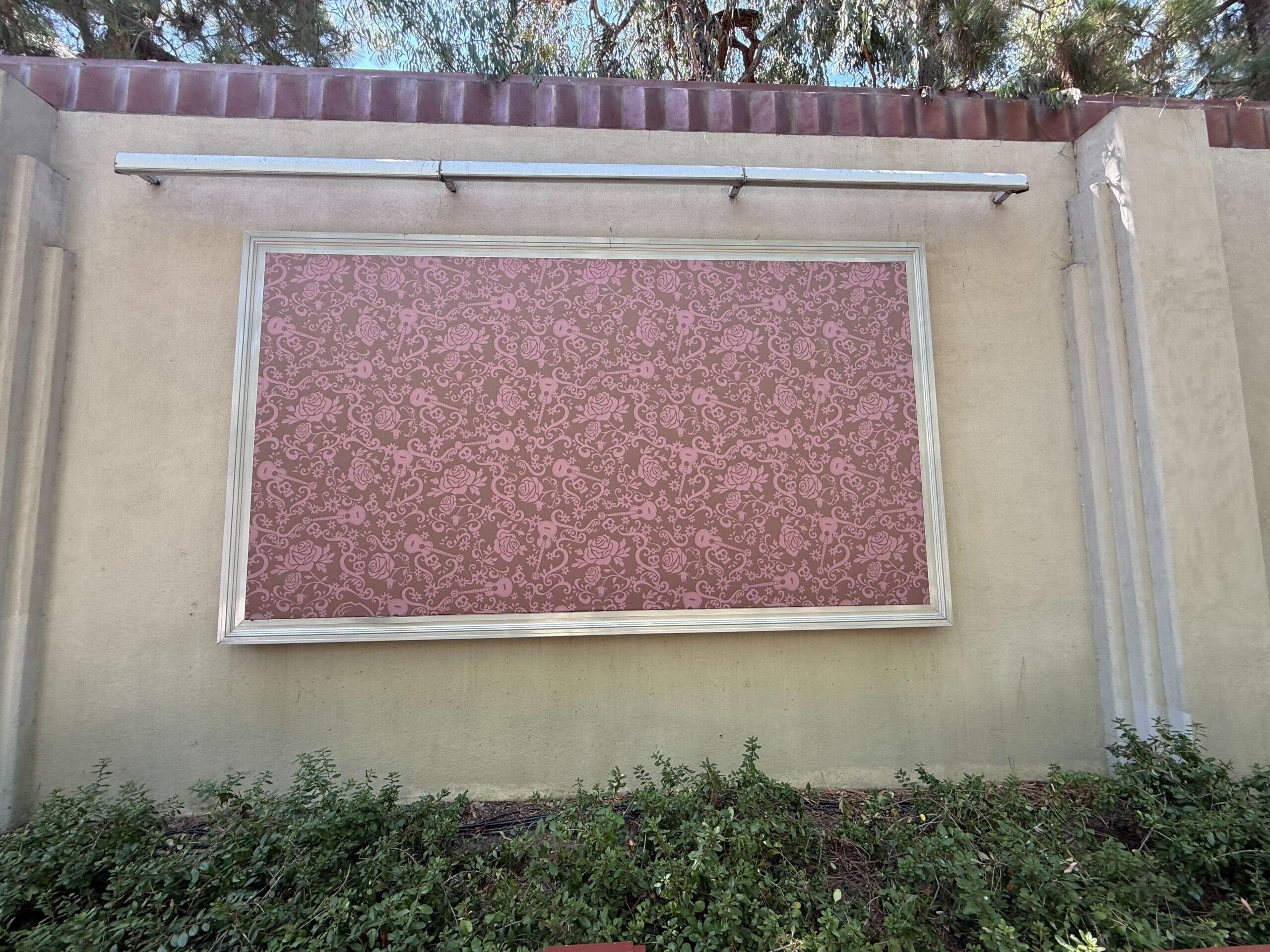 A large empty framed backdrop with a pink floral pattern, mounted on a beige wall above green shrubs.